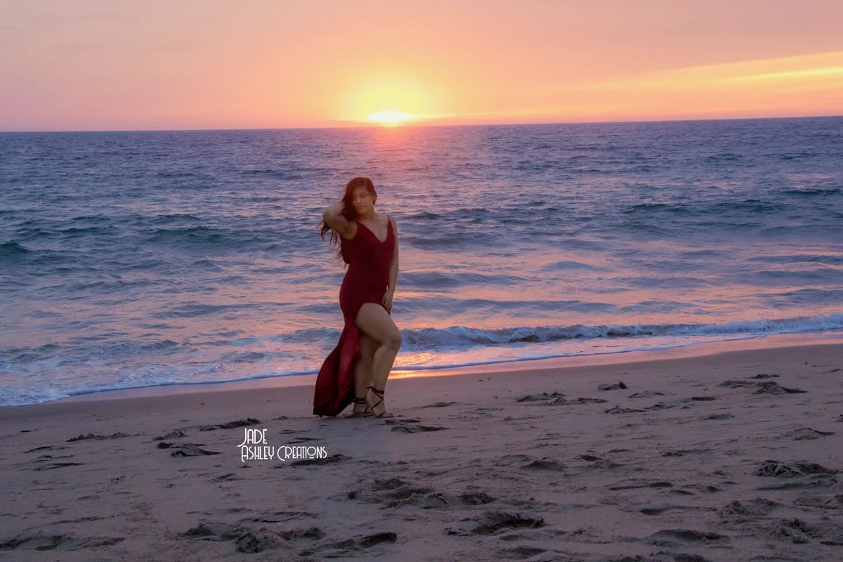 A woman in a red dress with a high slit standing on a beach at sunset, with the ocean and colorful sky in the background.