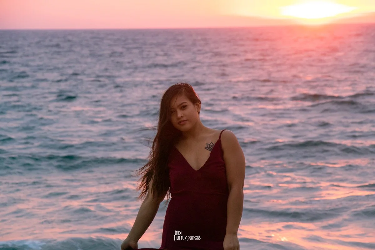 A woman with long hair in a maroon dress standing on the beach during sunset with the ocean in the background.