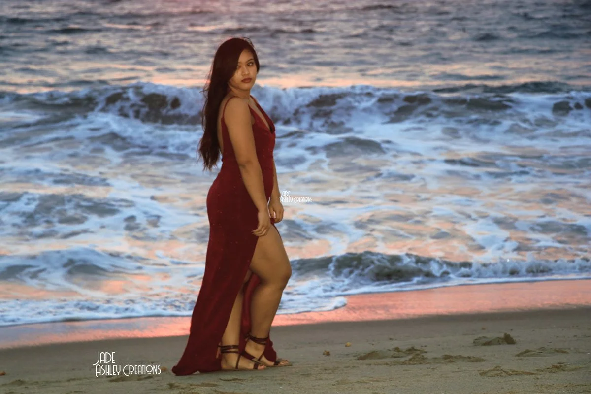 A woman in a red dress standing on a beach near the ocean at sunset.