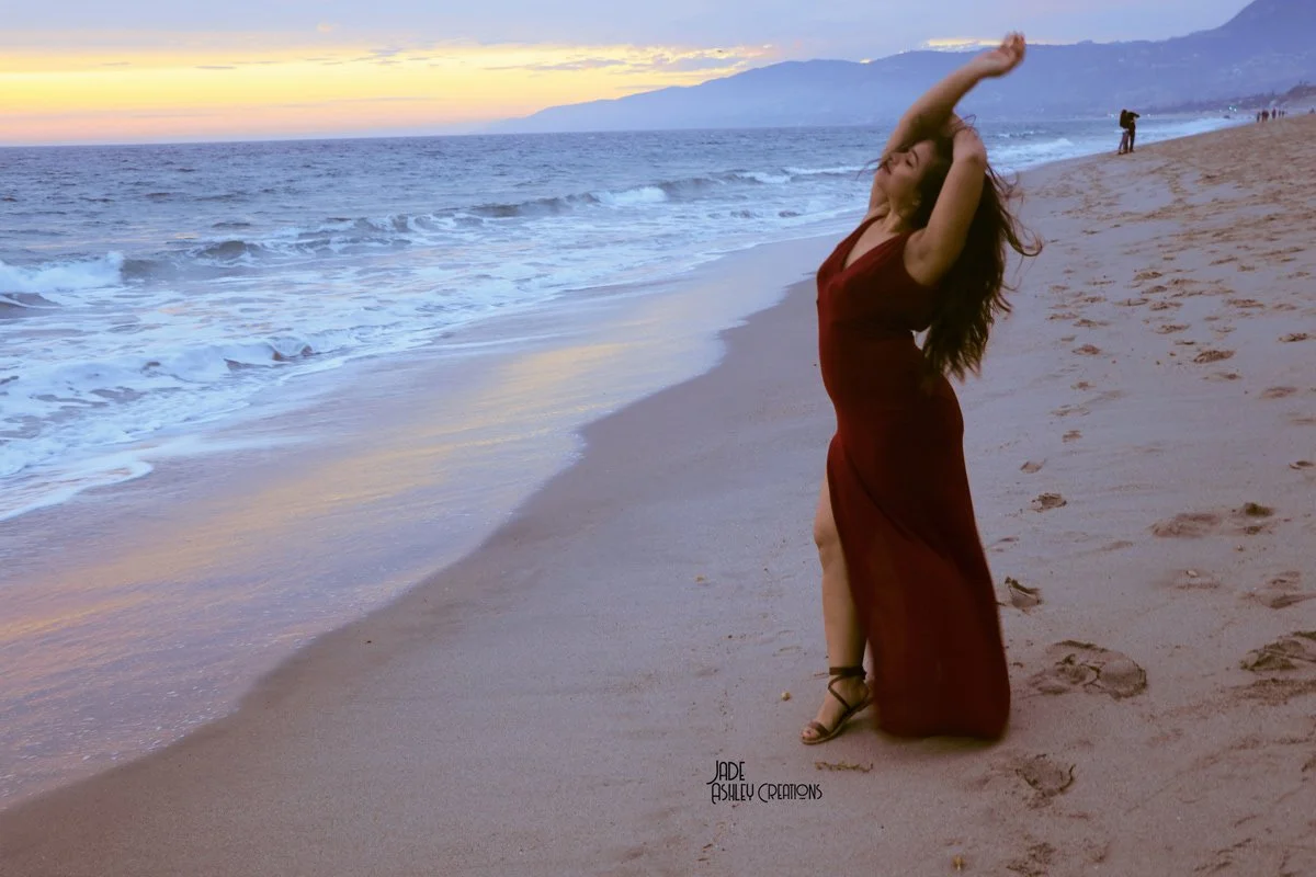 A woman in a red dress standing on a sandy beach at sunset, with mountains in the background and ocean waves nearby.