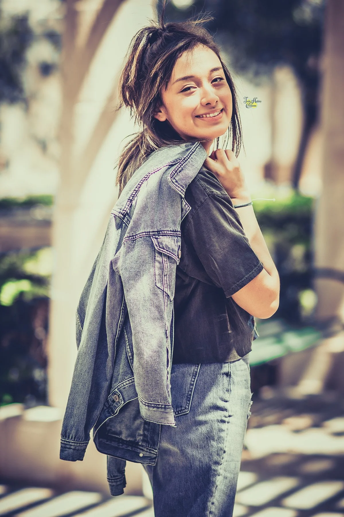 Young woman smiling, wearing a black t-shirt and jeans, holding a denim jacket over her shoulder, outdoors on a sunny day.