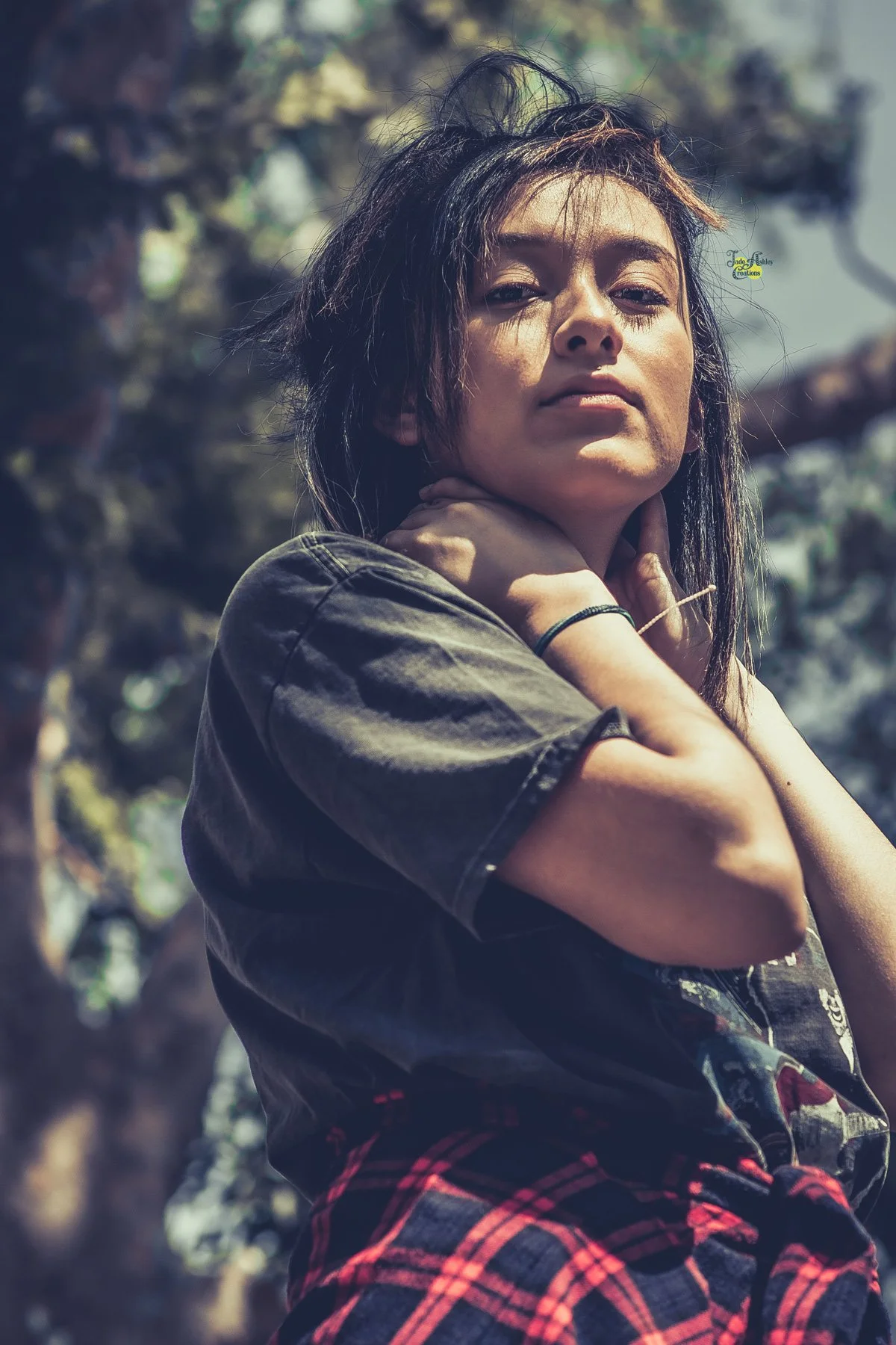 A young woman with long dark hair, wearing a black t-shirt and red plaid pants, standing outdoors with trees in the background.
