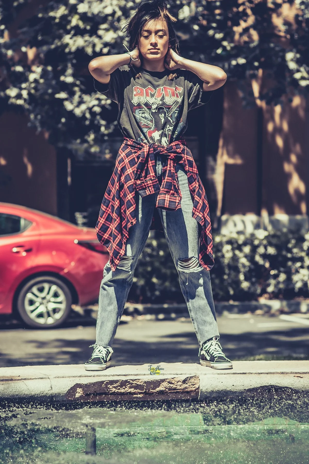 A young woman standing on a curb near a fountain, wearing a black AC/DC T-shirt, ripped jeans, and sneakers with a red and black plaid shirt tied around her waist. She has her hands behind her neck and is looking downward, with trees and a red car in