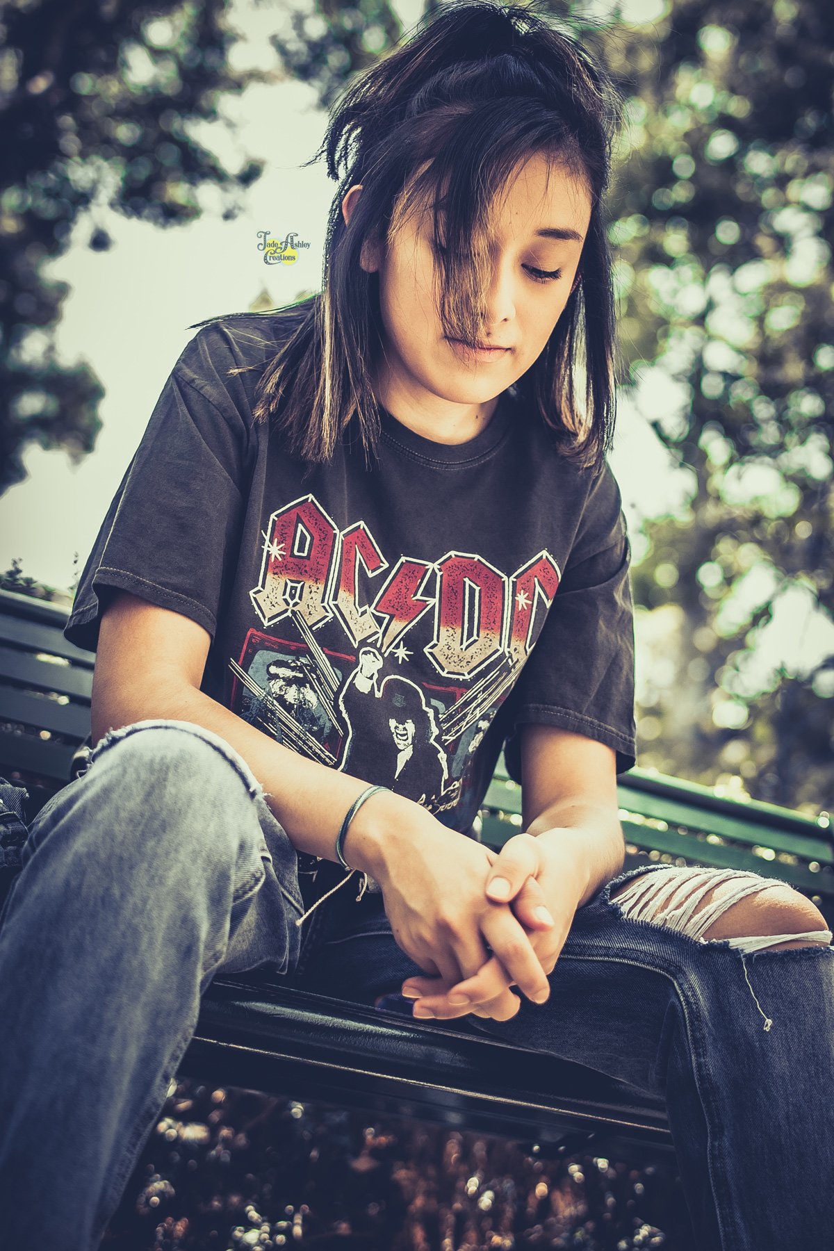 A young woman with black hair sitting on a park bench, wearing a black AC/DC band t-shirt and ripped jeans, looking down with her hands clasped in her lap.
