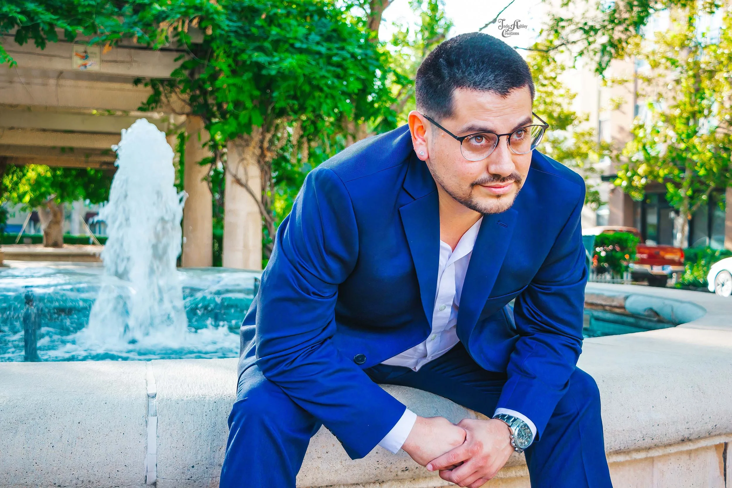 A man wearing a blue suit and glasses sitting on a stone bench by a fountain in an outdoor park area with trees and buildings in the background.