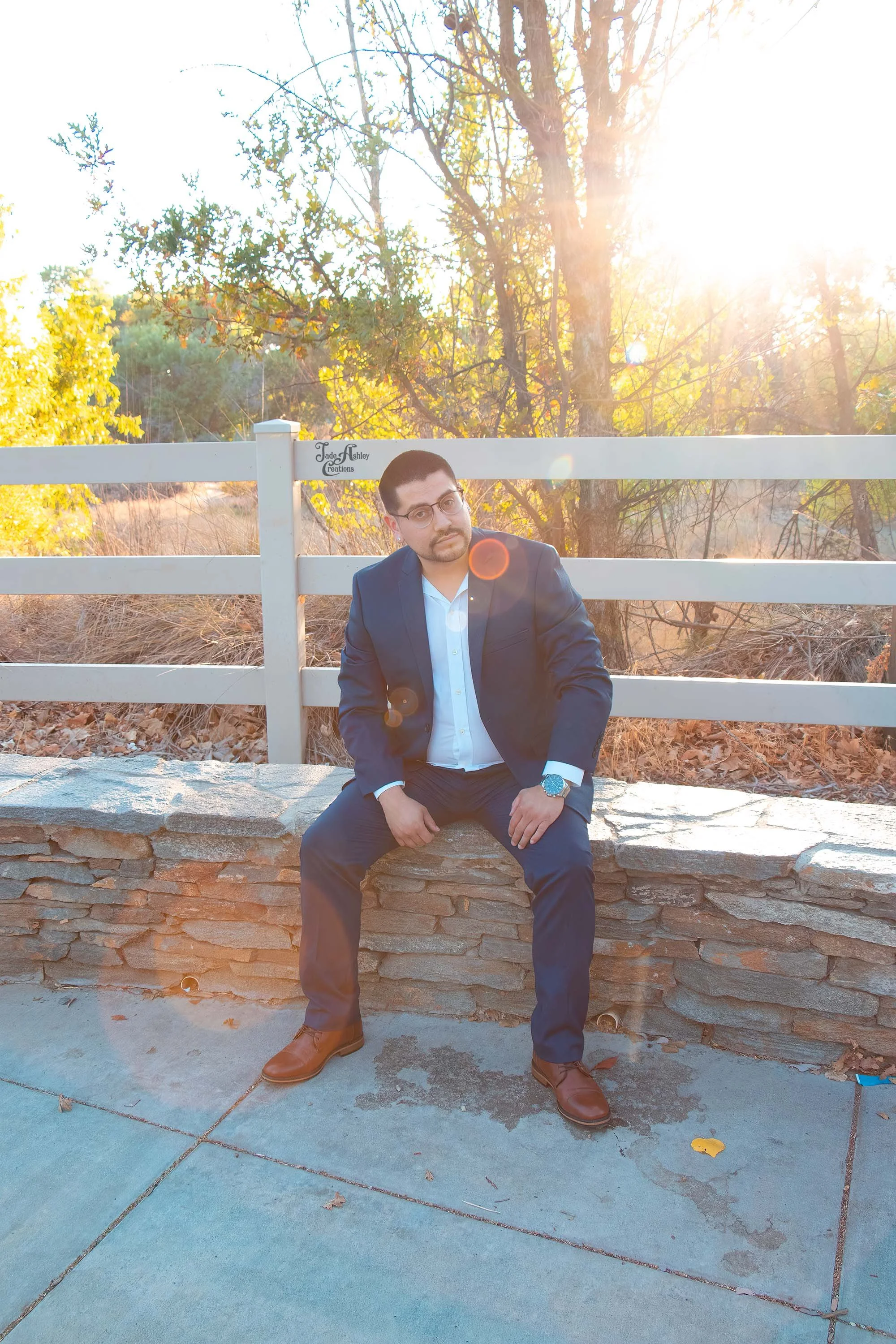 A man in a blue suit and white shirt sitting on a stone ledge outdoors with a white fence behind him, trees in the background, and sunlight shining through the branches.