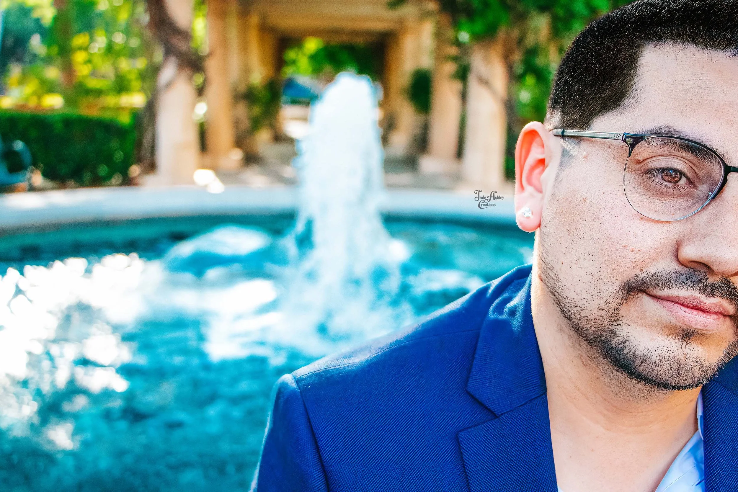 Partial close-up of a man with dark hair, glasses, and a beard, wearing a blue blazer and a white shirt, standing outdoors with a fountain and trees in the background.