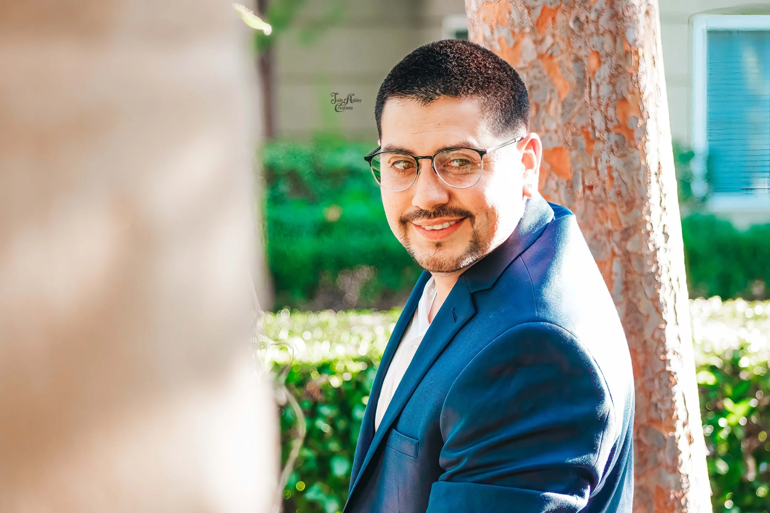 A man with short dark hair, glasses, and a goatee, smiling while wearing a blue suit jacket and a white shirt, standing outdoors beside a tree.