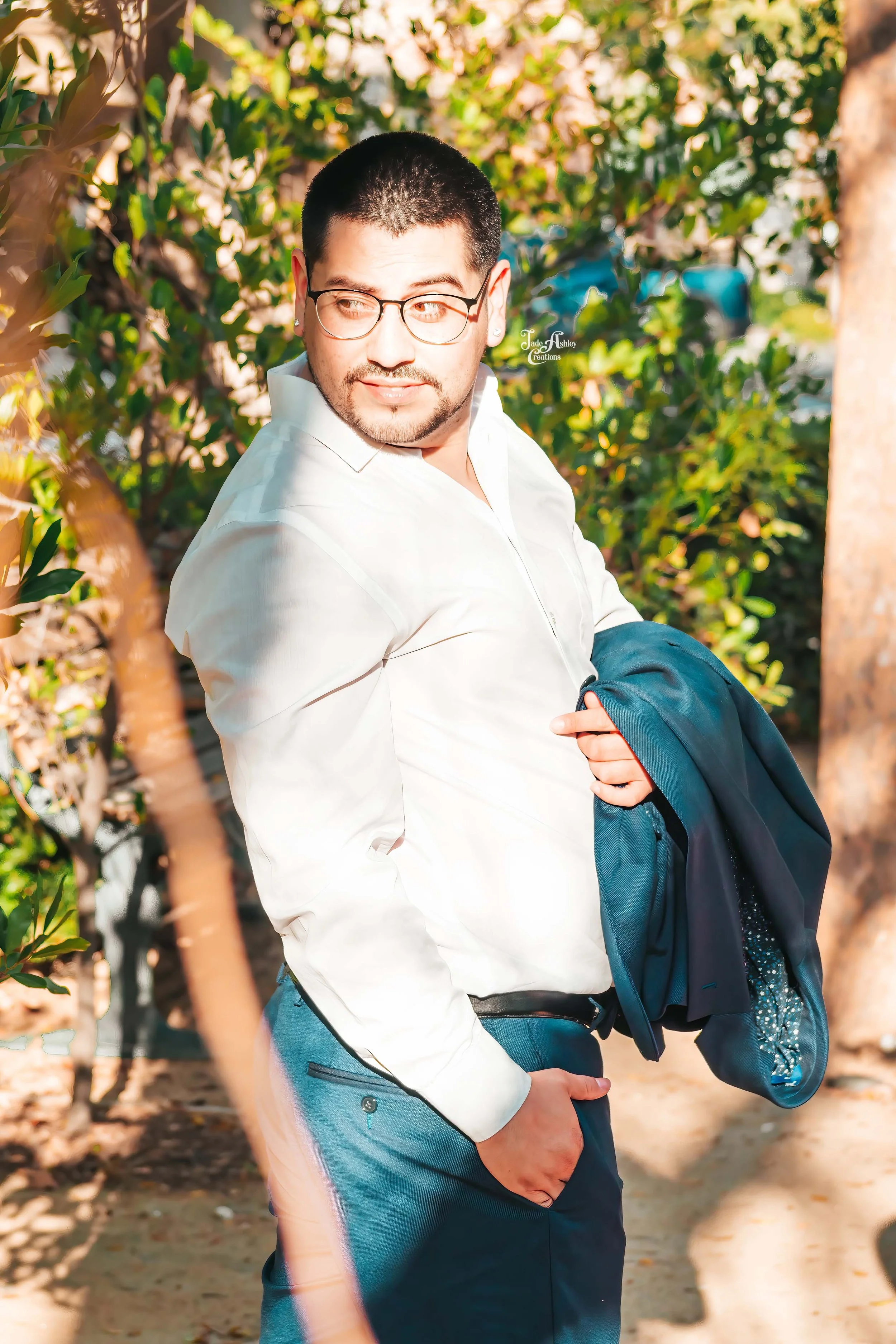 A man in a white shirt holding a dark suit jacket and standing outdoors among greenery, looking to the side.