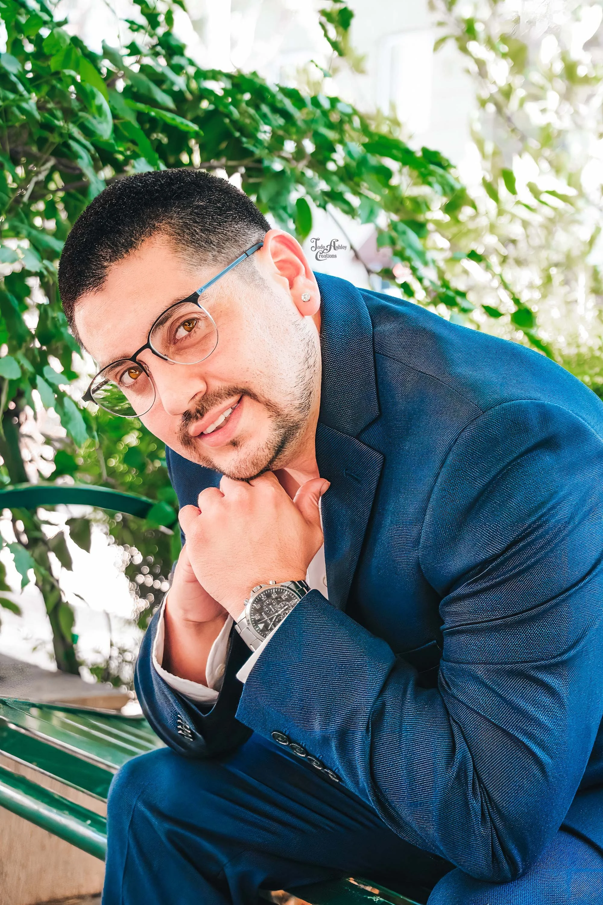 A man with short dark hair, glasses, and a beard, wearing a navy blue suit and watch, is sitting on a green bench outdoors with lush green foliage in the background, resting his chin on clasped hands and smiling at the camera.