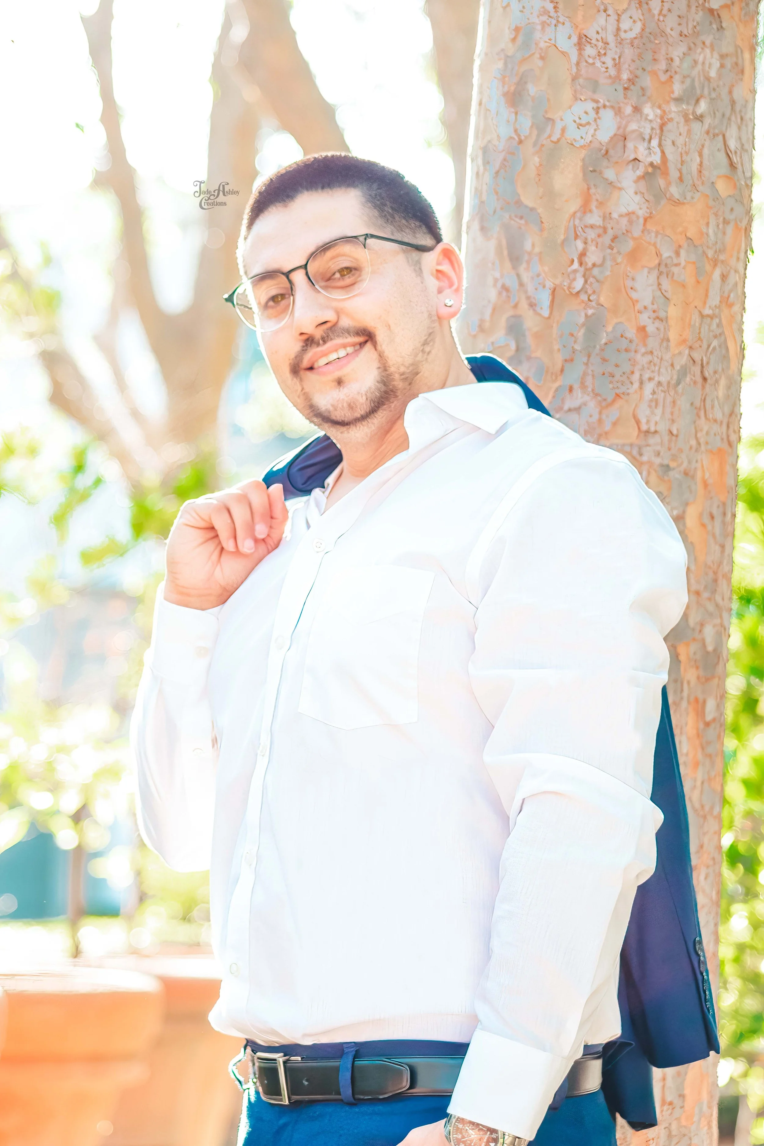 A young man with short dark hair, glasses, and earrings, wearing a white shirt and dark pants, stands outdoors beside a tree. He is smiling and carrying a dark jacket over his shoulder. The background features bright sunlight, greenery, and a blurred