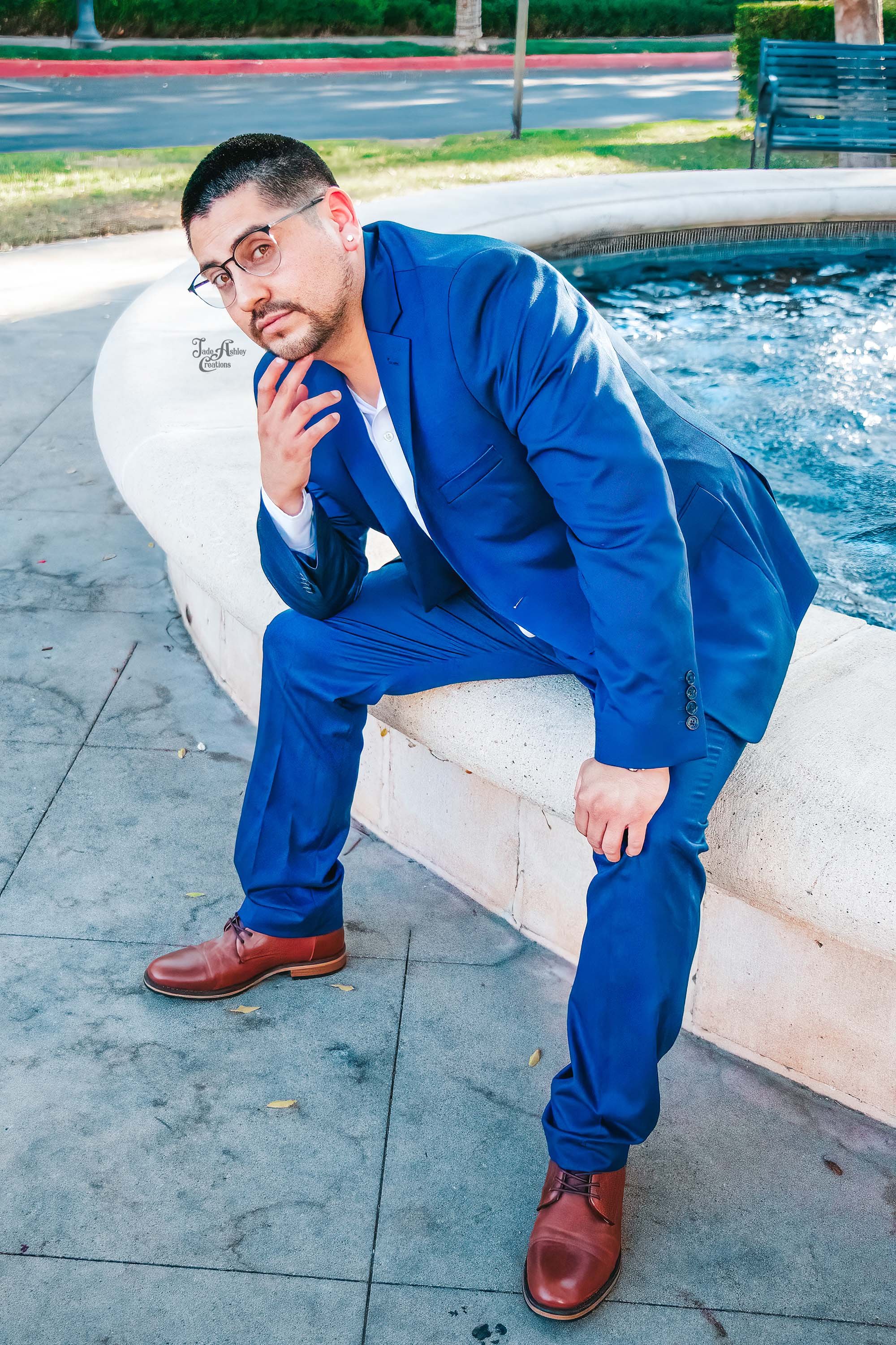 A man in a blue suit and brown boots sitting on the edge of a fountain outdoors, with trees, a bench, and a road in the background.