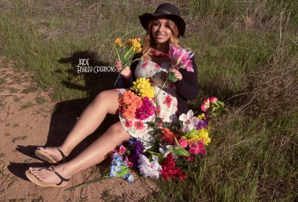 Woman sitting on dirt ground surrounded by colorful flowers, wearing a floral dress, black cardigan, sun hat, and sandals, holding yellow and pink flowers