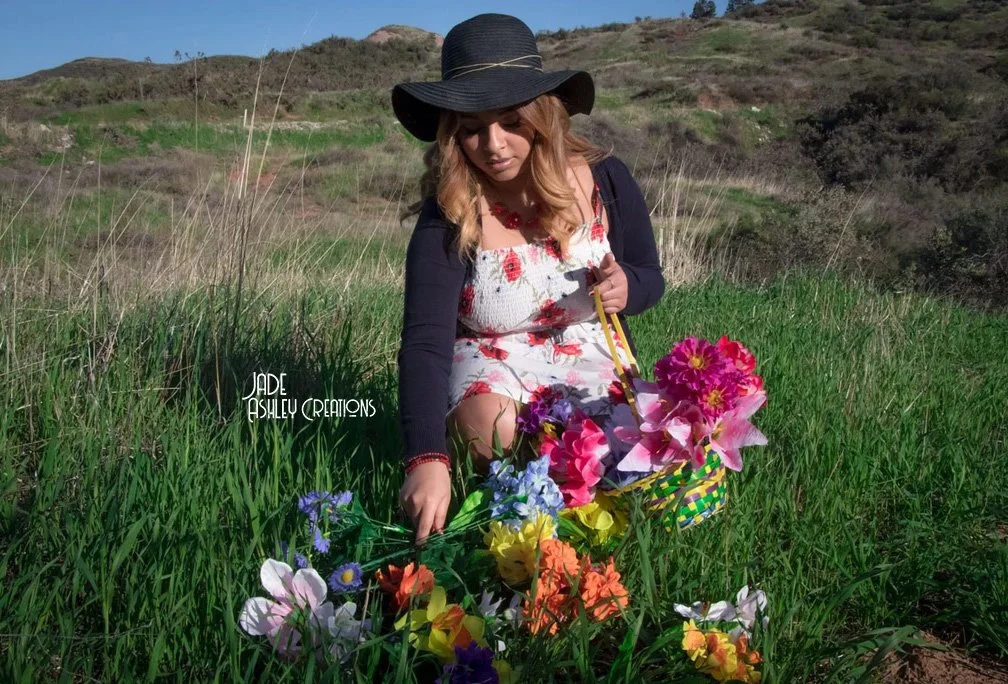 A woman wearing a wide-brimmed black hat and a floral dress with a black cardigan is sitting in a grassy field with hills in the background. She is surrounded by colorful paper flowers in a basket, suggesting an outdoor spring or summer setting.