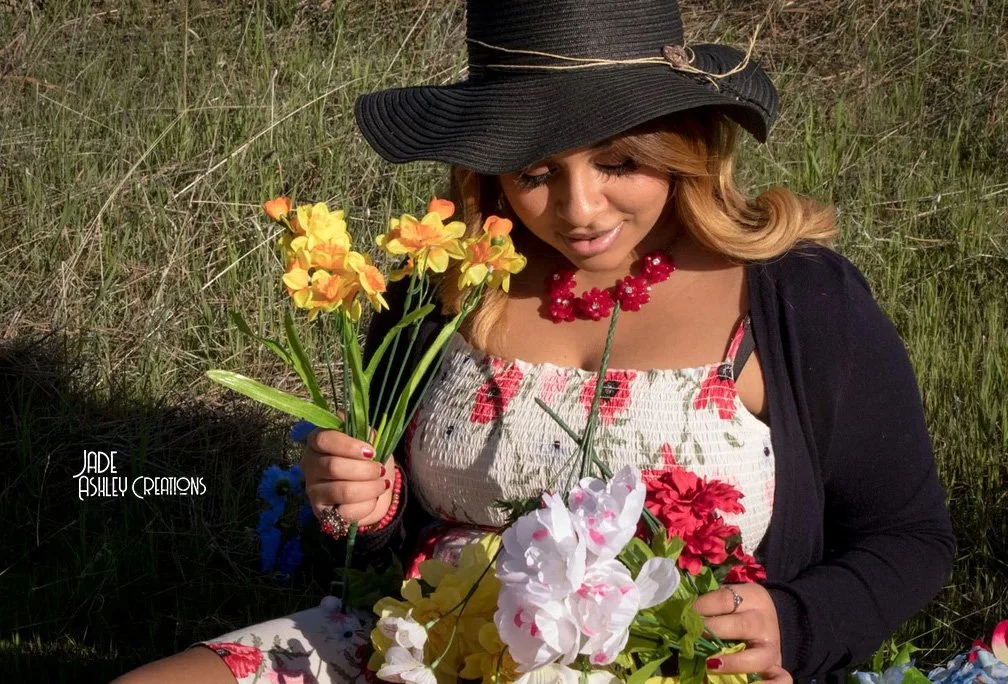 Woman wearing a large black sun hat and floral dress holds colorful artificial flowers outdoors with grassy background.