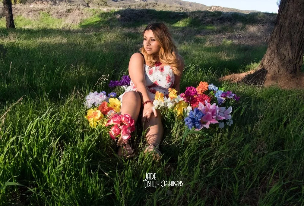 A woman sitting in green grass surrounded by colorful artificial flowers in an outdoor setting with trees and hills in the background.