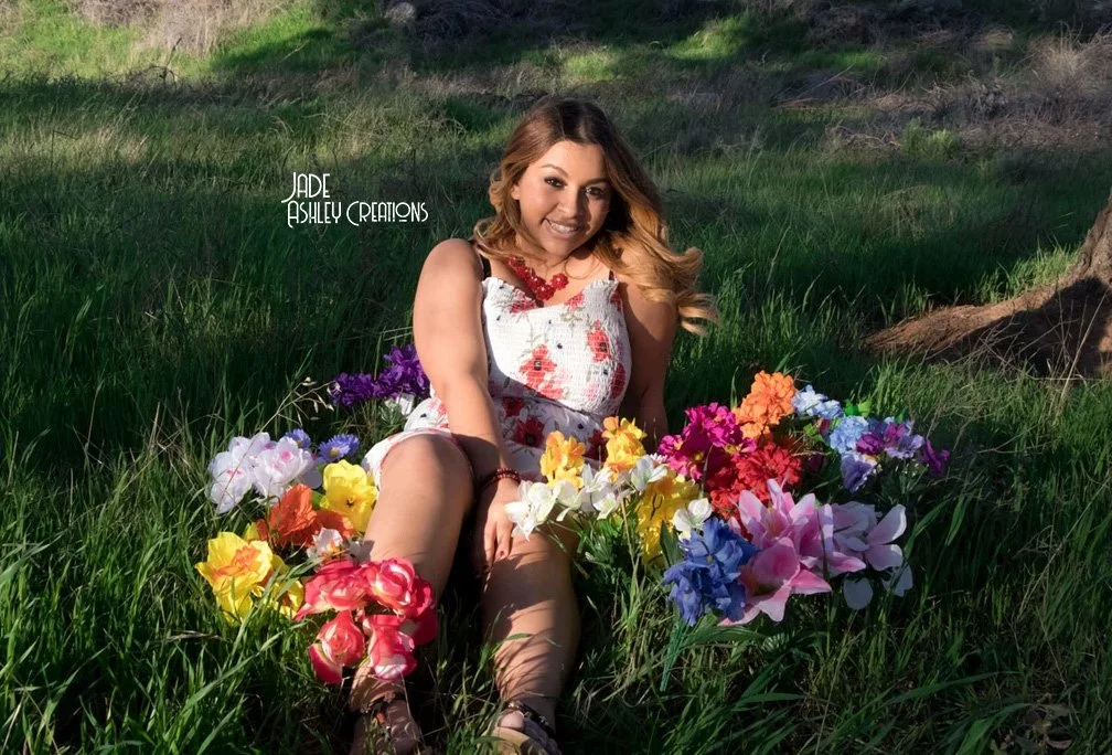A woman sitting on the grass surrounded by colorful flowers, wearing a white dress with red floral patterns, with long wavy hair and a necklace, in a natural outdoor setting.