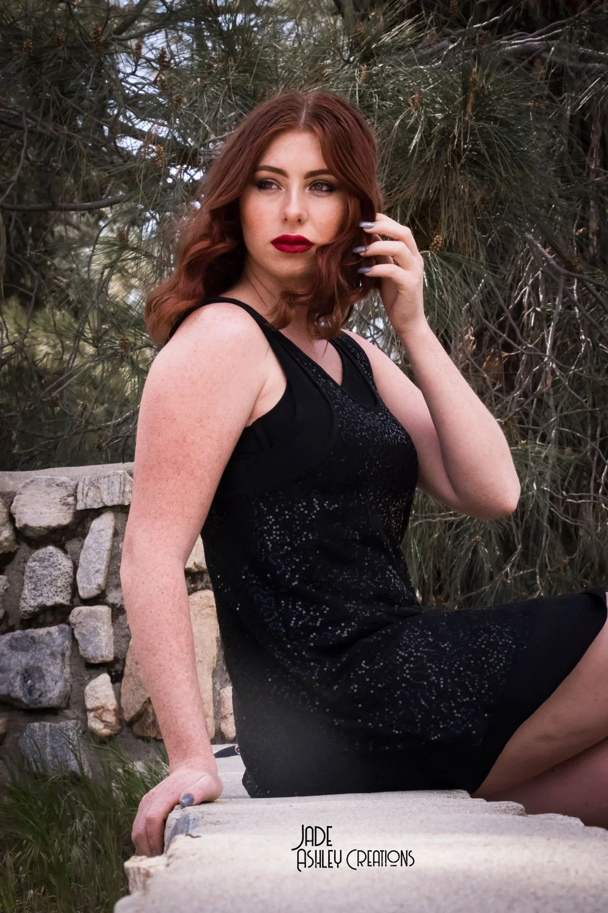 A woman with wavy red hair, wearing a black sleeveless dress with sparkles, sitting outdoors on a stone ledge with greenery and trees in the background.