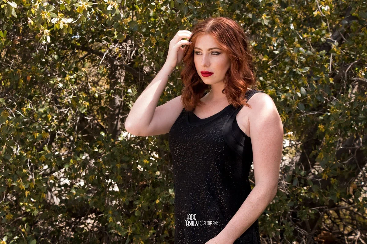 A woman with red hair, wearing a black dress, stands outdoors in front of green bushes, touching her hair with one hand.