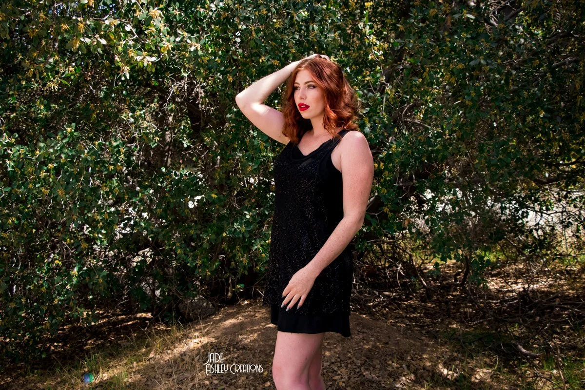 A woman with red hair and red lipstick standing outdoors in front of green bushes, wearing a sleeveless black dress.