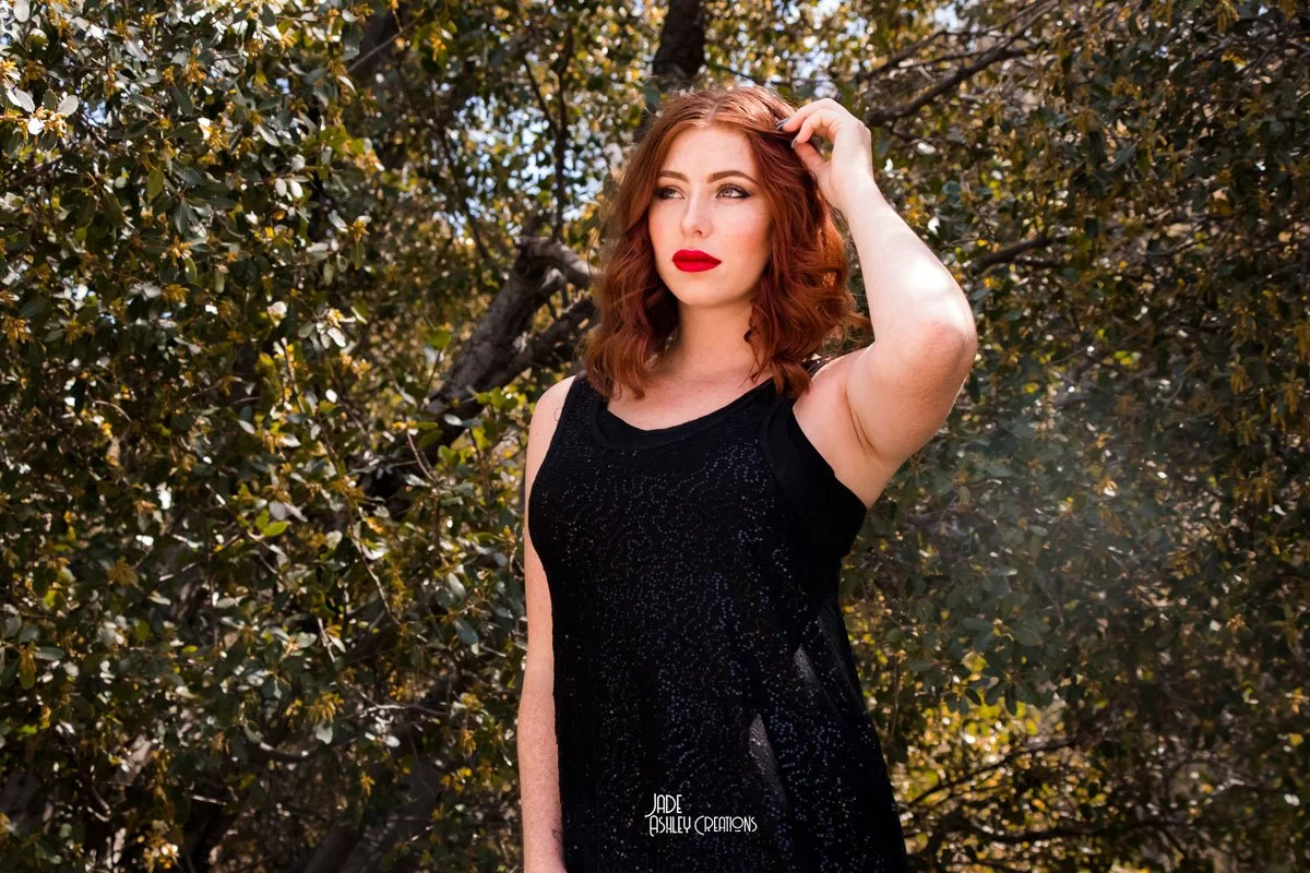 A woman with red hair and red lipstick wearing a sleeveless black dress standing outdoors in front of leafy trees.
