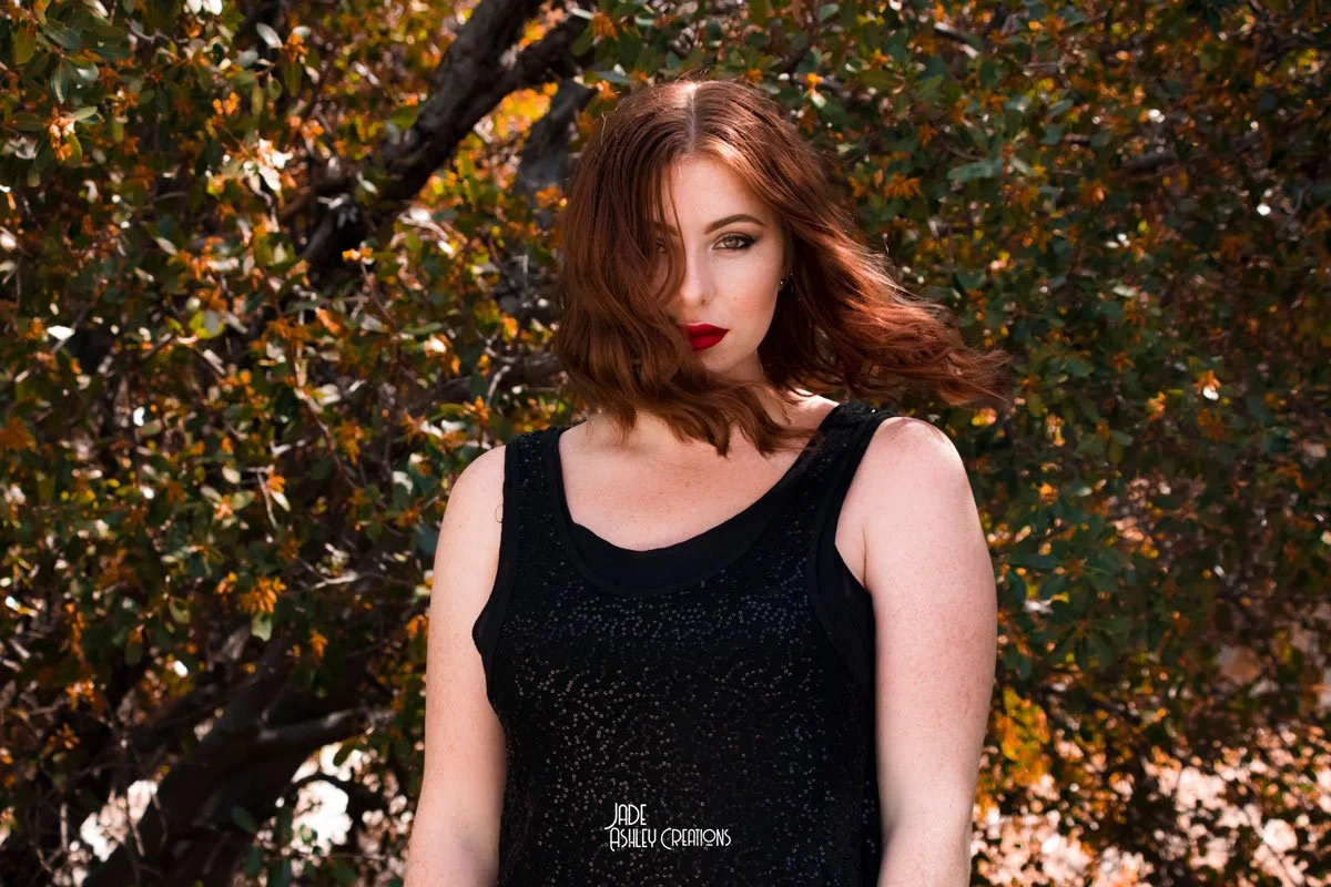 A young woman with red hair, red lipstick, and no shirt visible, standing outdoors in front of a background of trees with green and yellow leaves.