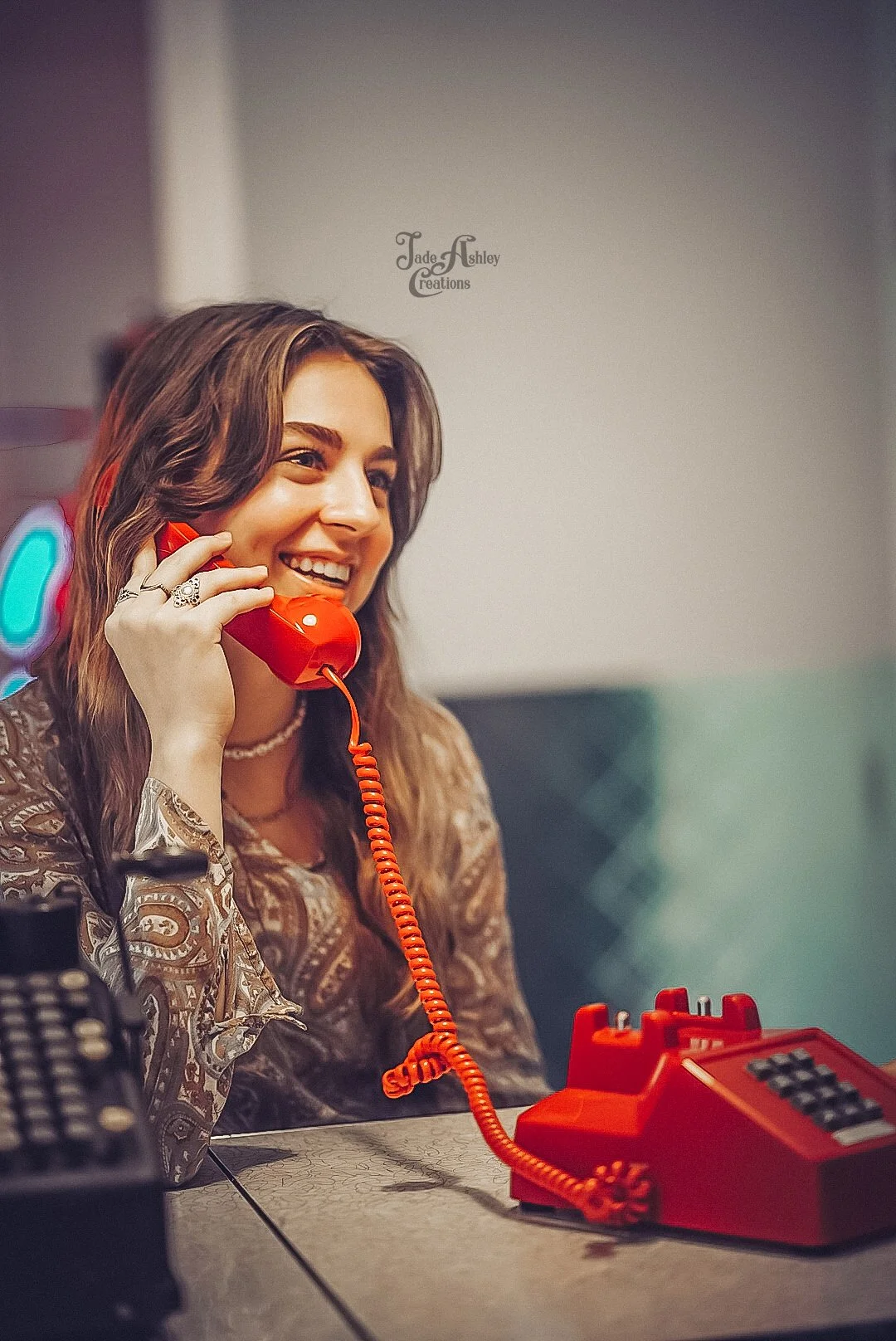A woman with wavy brown hair, wearing jewelry and a decorative top, smiling while talking on a red rotary phone at a desk in a retro style setting.