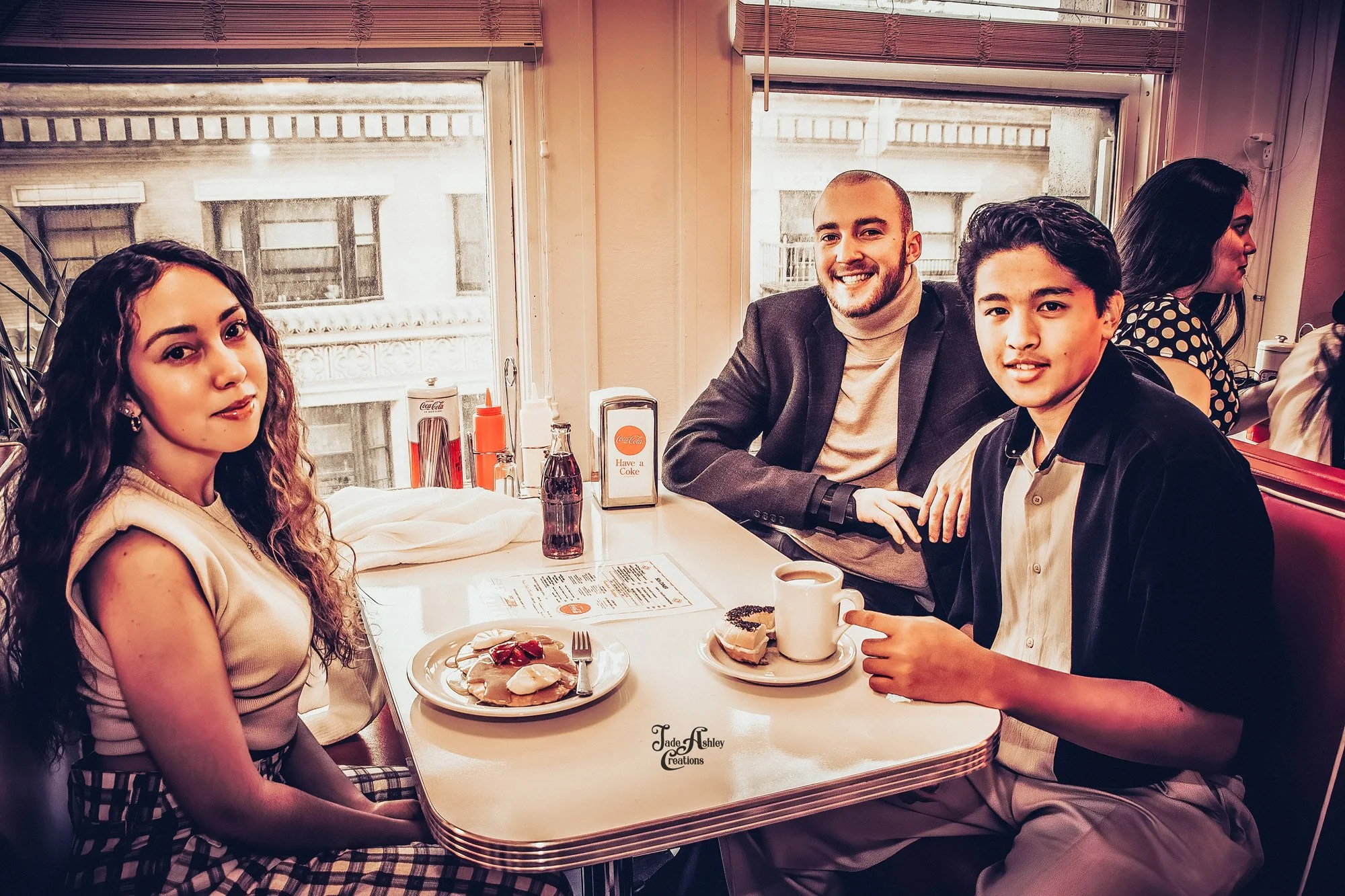 Group of four young people sitting at a diner table with food and drinks, smiling at the camera, in a vintage-style restaurant with large windows.