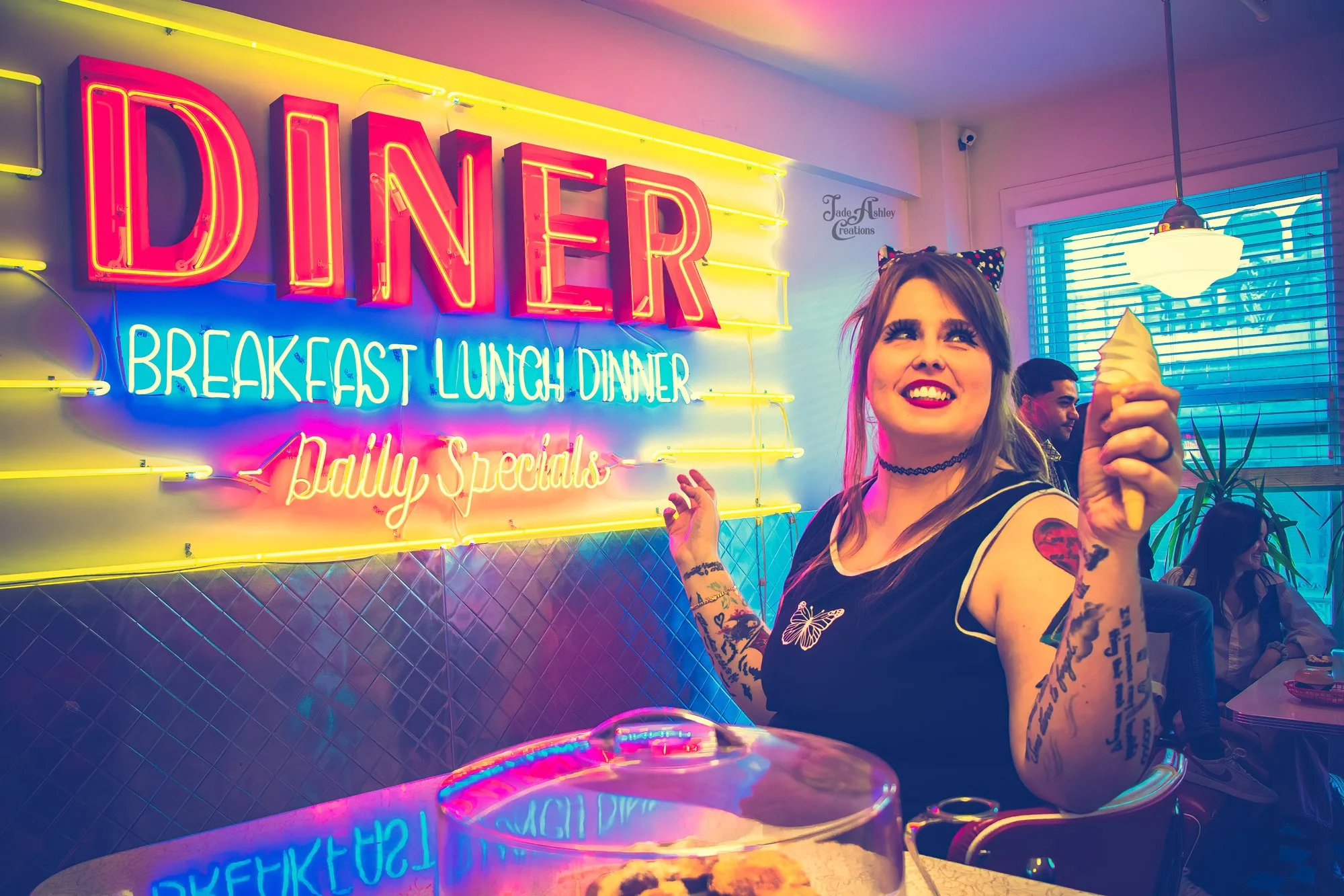 A woman with tattoos, wearing a butterfly patch and cat ear headband, smiling and holding an ice cream cone inside a colorful neon-lit diner with a vibrant sign in the background that reads 'Diner' and includes breakfast, lunch, dinner, and daily spe