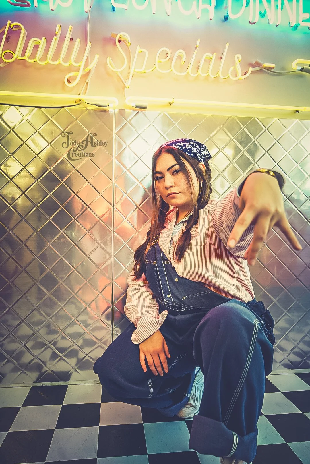 Young woman with braids and a bandana posing in front of a neon sign reading 'Daily Specials.' She is dressed in overalls and a striped shirt, squatting on a checkered floor, reaching out with her right hand and looking at the camera.