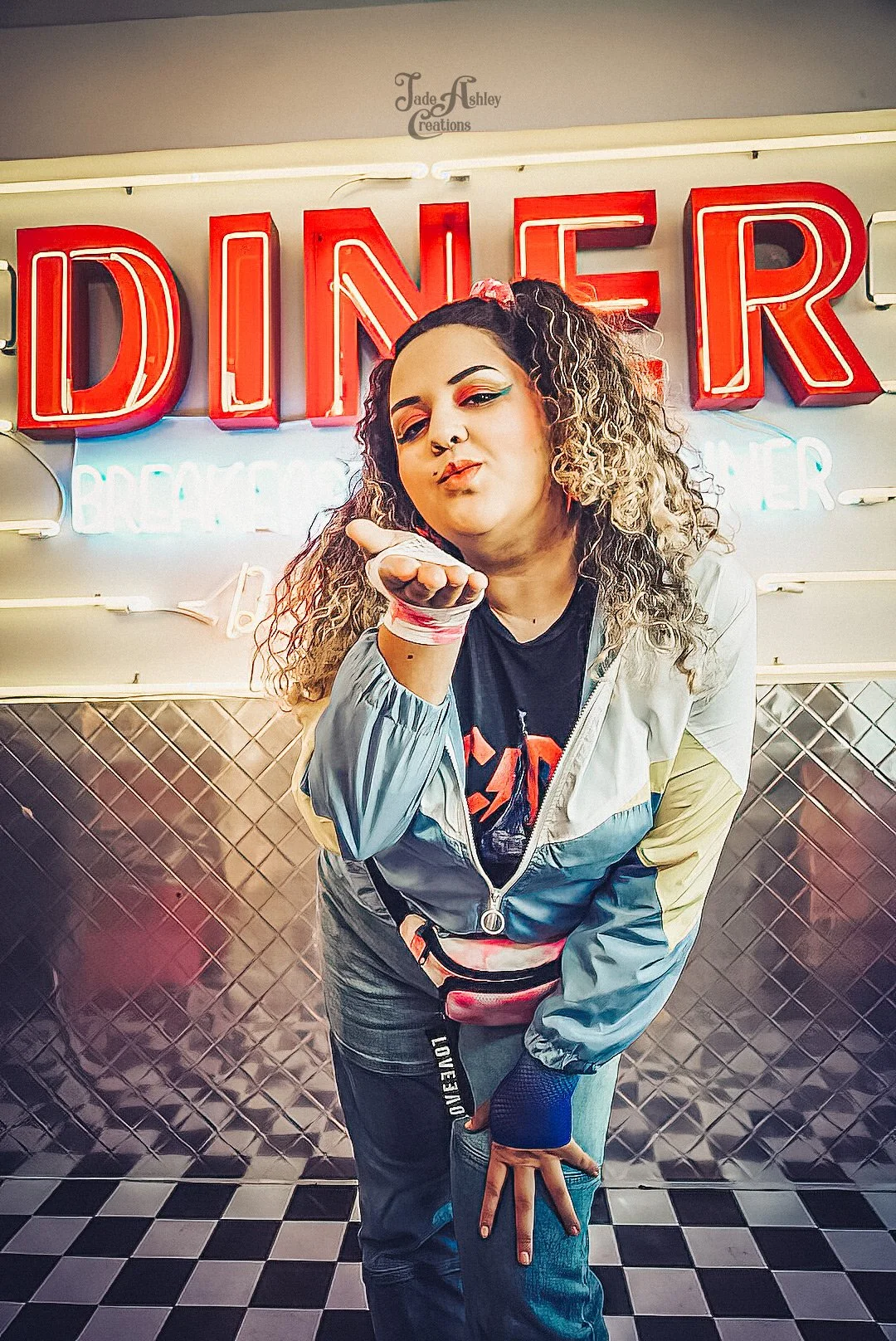 A woman with curly hair and makeup blowing a kiss in front of a neon sign that says 'DINER'. She is wearing a color-block jacket and jeans, with a pink fanny pack. The background has a checkered floor pattern.