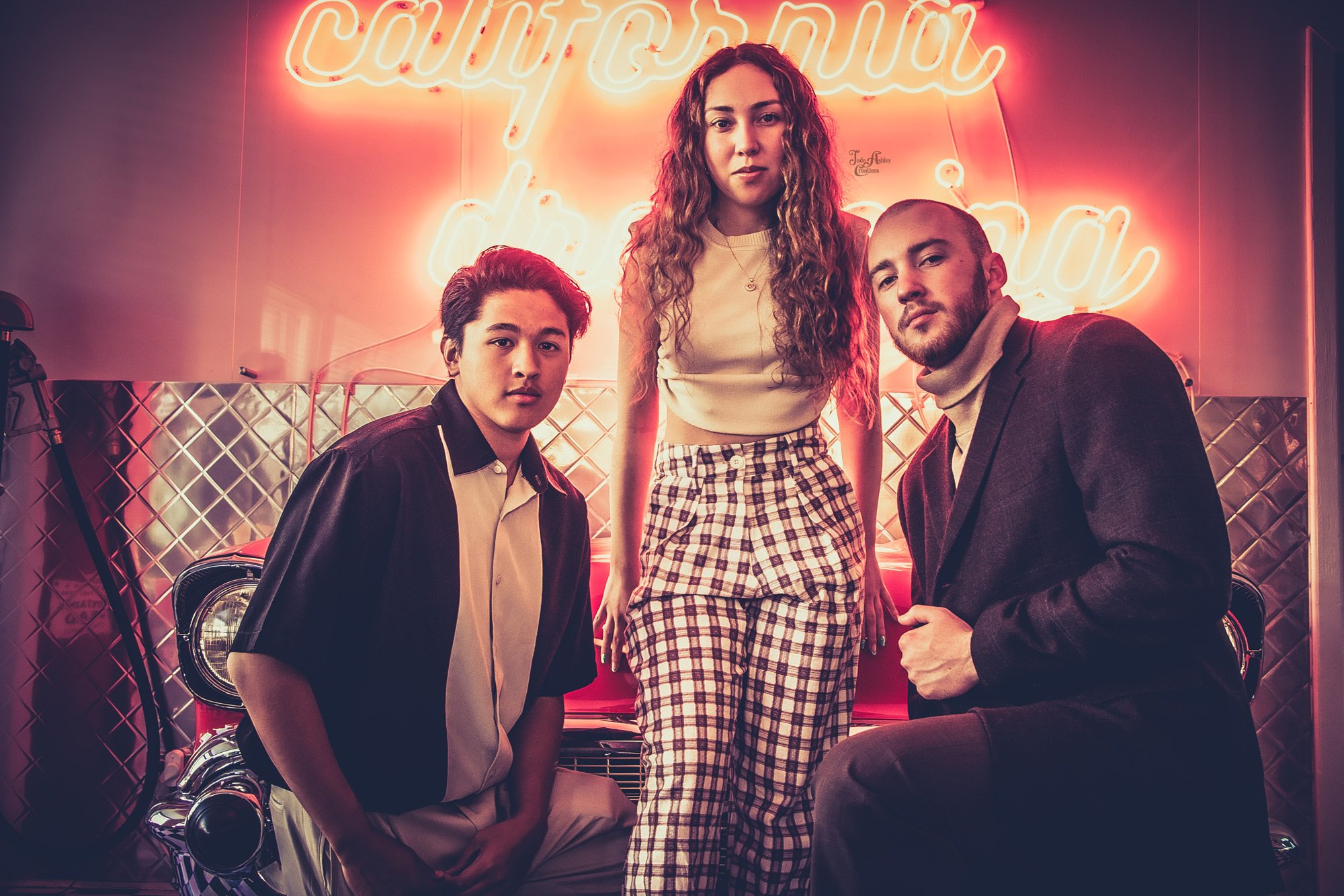 A group of three young adults, two men and one woman, posing in front of a neon sign that reads 'caffeine over cardio.' The setting has a vintage vibe with a classic car behind them and a metallic wall. The woman has long curly hair and wears plaid p