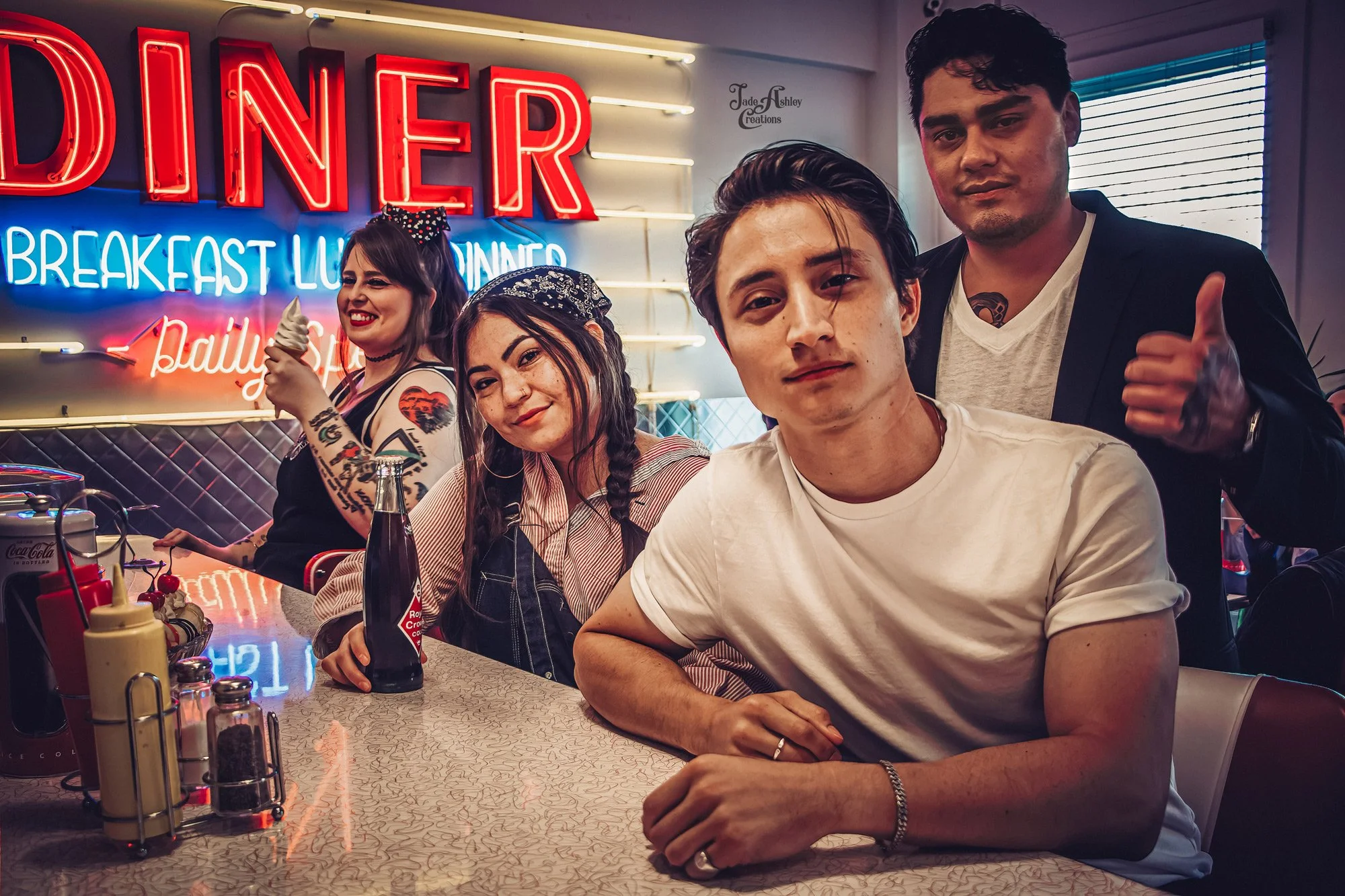 Four young adults sitting at a diner counter with neon signs in the background, smiling and posing for the photo.