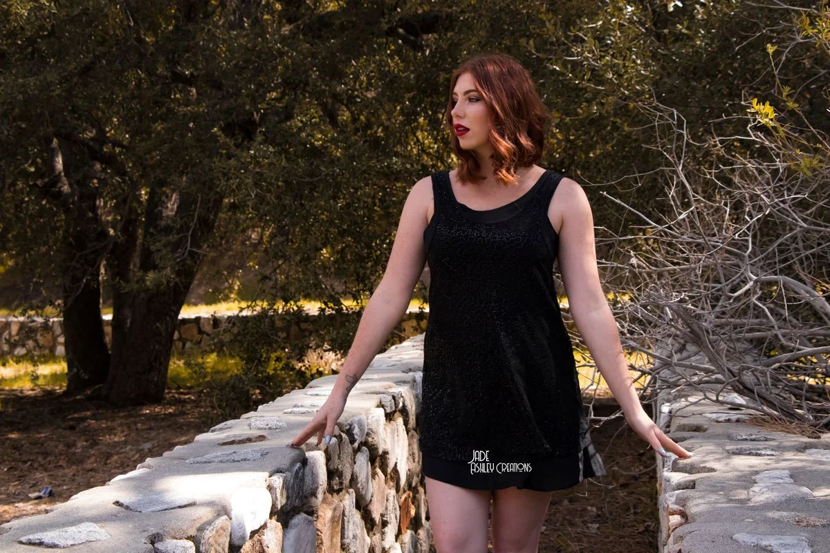 A young woman with shoulder-length red hair and fair skin, wearing a sleeveless black dress, stands on a stone bridge surrounded by trees and natural scenery.