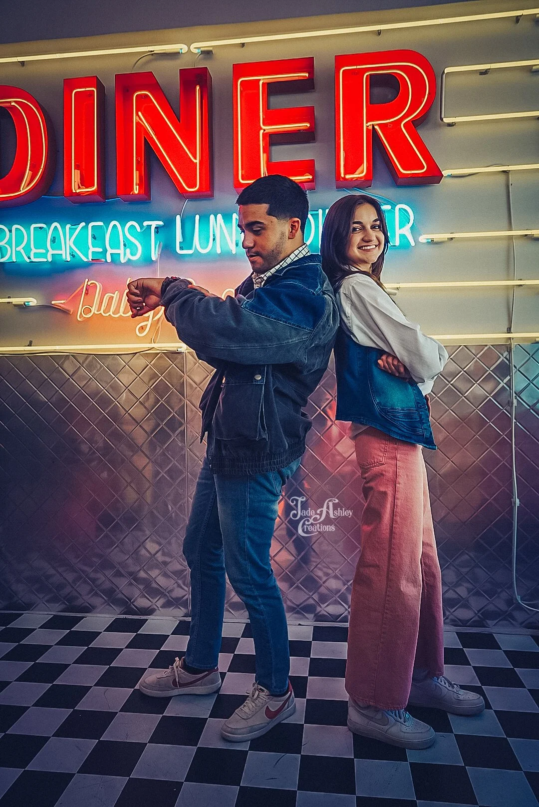 Two young people standing back-to-back in front of a neon diner sign, one checking their watch and the other smiling. The sign says 'DINER' and lists breakfast, lunch, and dinner options, with a checkered black and white floor.