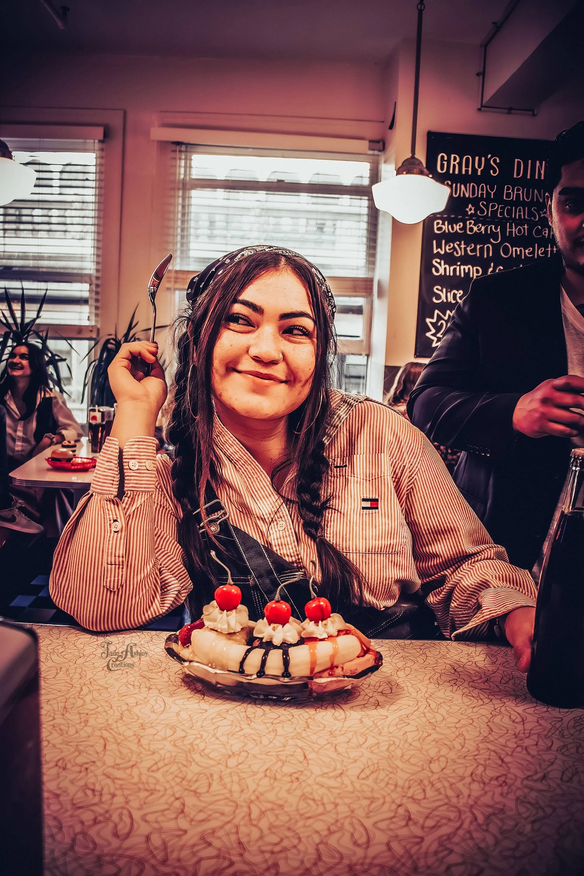 A woman with long braided hair smiling at the camera, sitting at a table with a banana split topped with whipped cream, cherry, and chocolate drizzle. She is wearing a striped shirt and headscarf. In the background, other people are in a diner-style 
