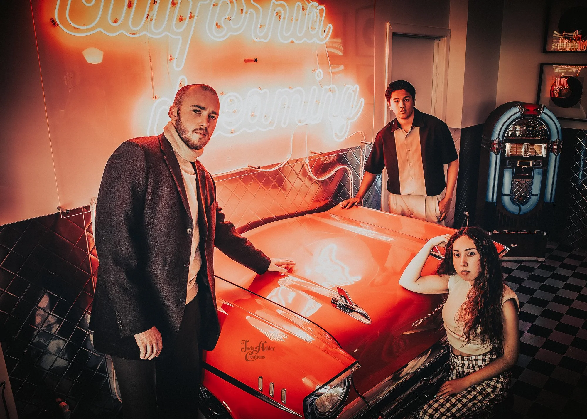 Four young adults posing around a red vintage car inside a retro-style room with neon lighting and a jukebox in the background.