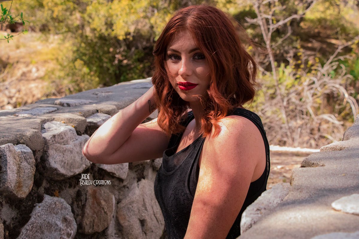 A woman with wavy red hair and red lipstick leans on a stone wall outdoors, wearing a black sleeveless top, with trees and foliage in the background.