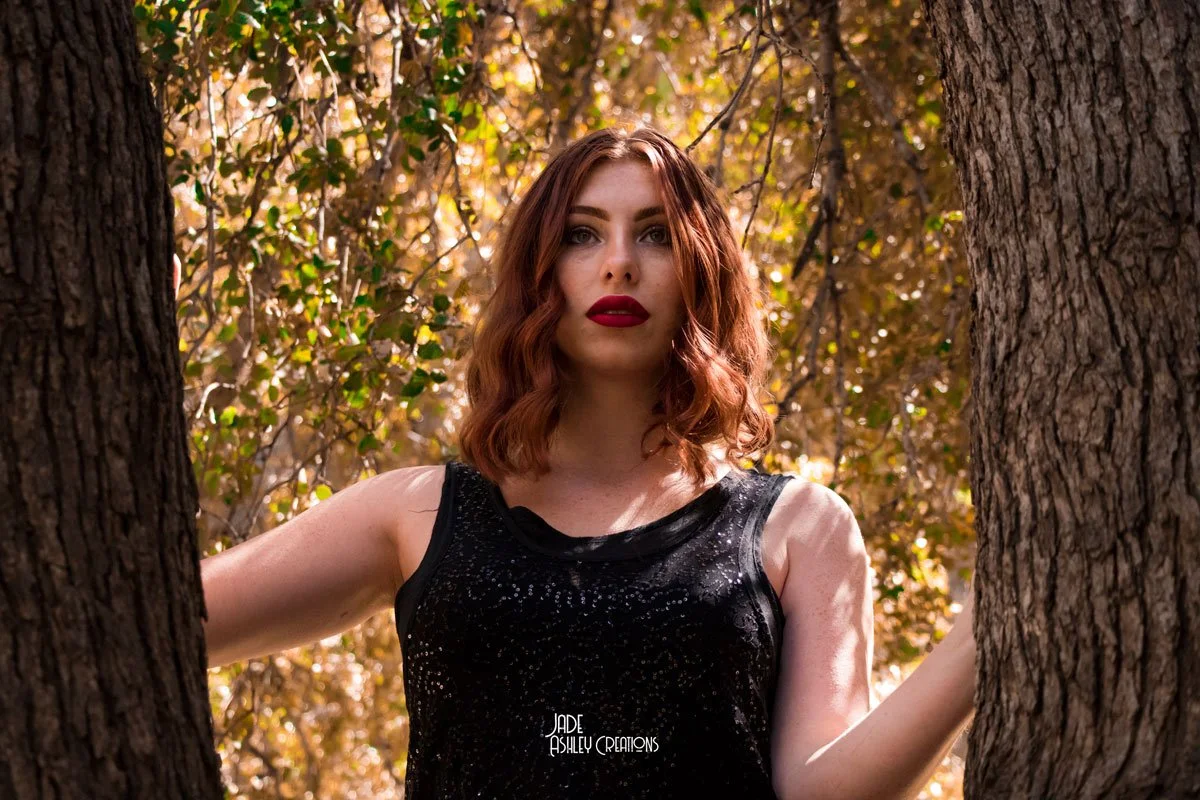 A young woman with curly auburn hair and red lipstick standing outdoors between two trees with autumn-colored leaves in the background.