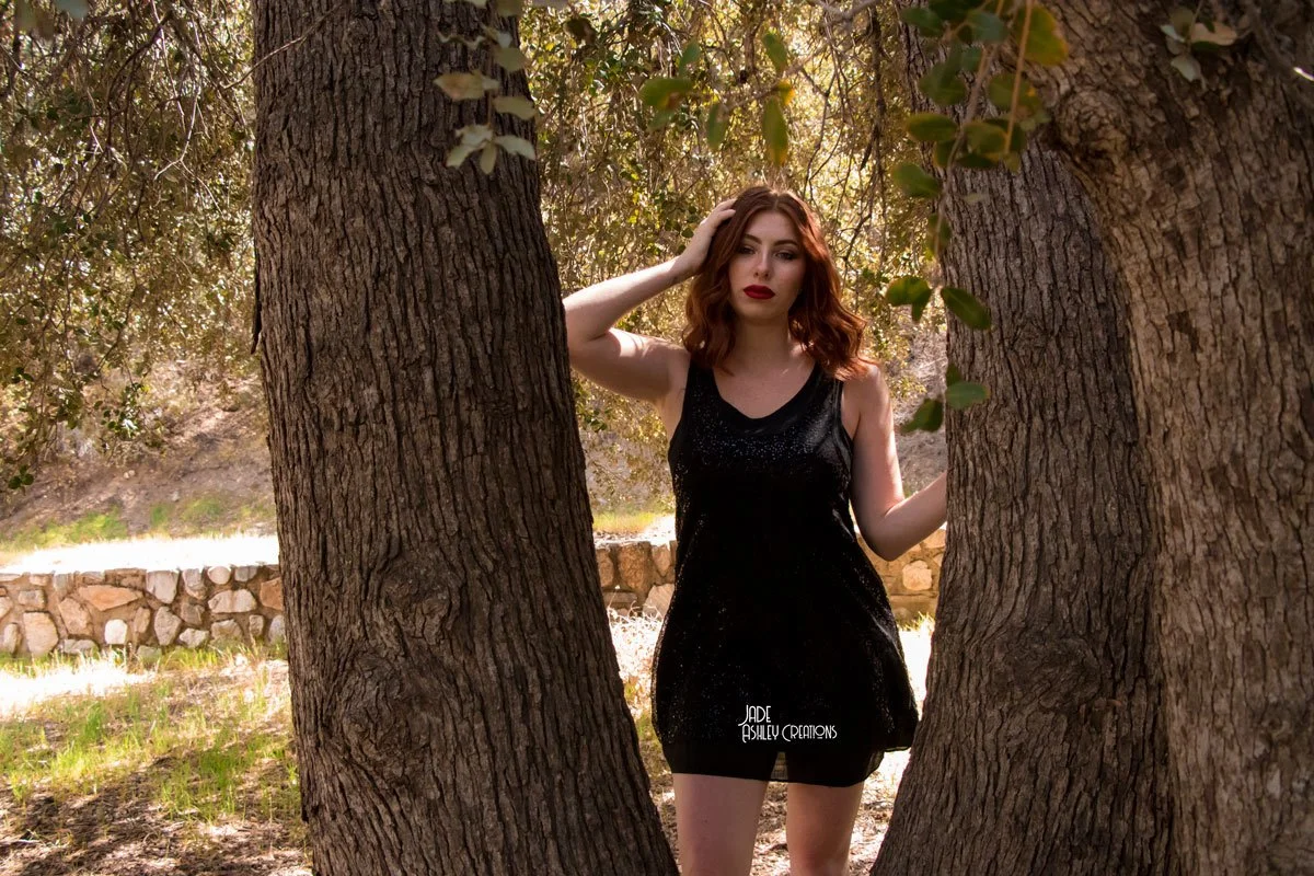 A young woman in a black dress standing between two large tree trunks in an outdoor setting with a stone wall in the background.