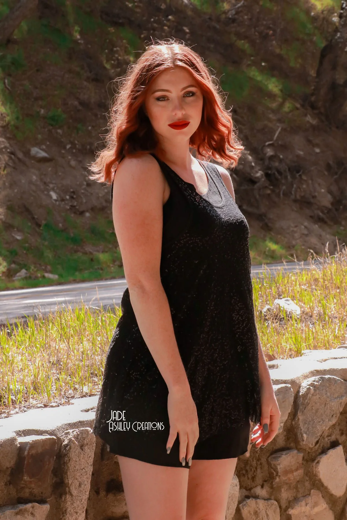 A young woman with red hair and fair skin standing outdoors by a stone ledge, wearing a black sleeveless dress, with a rocky hillside in the background.