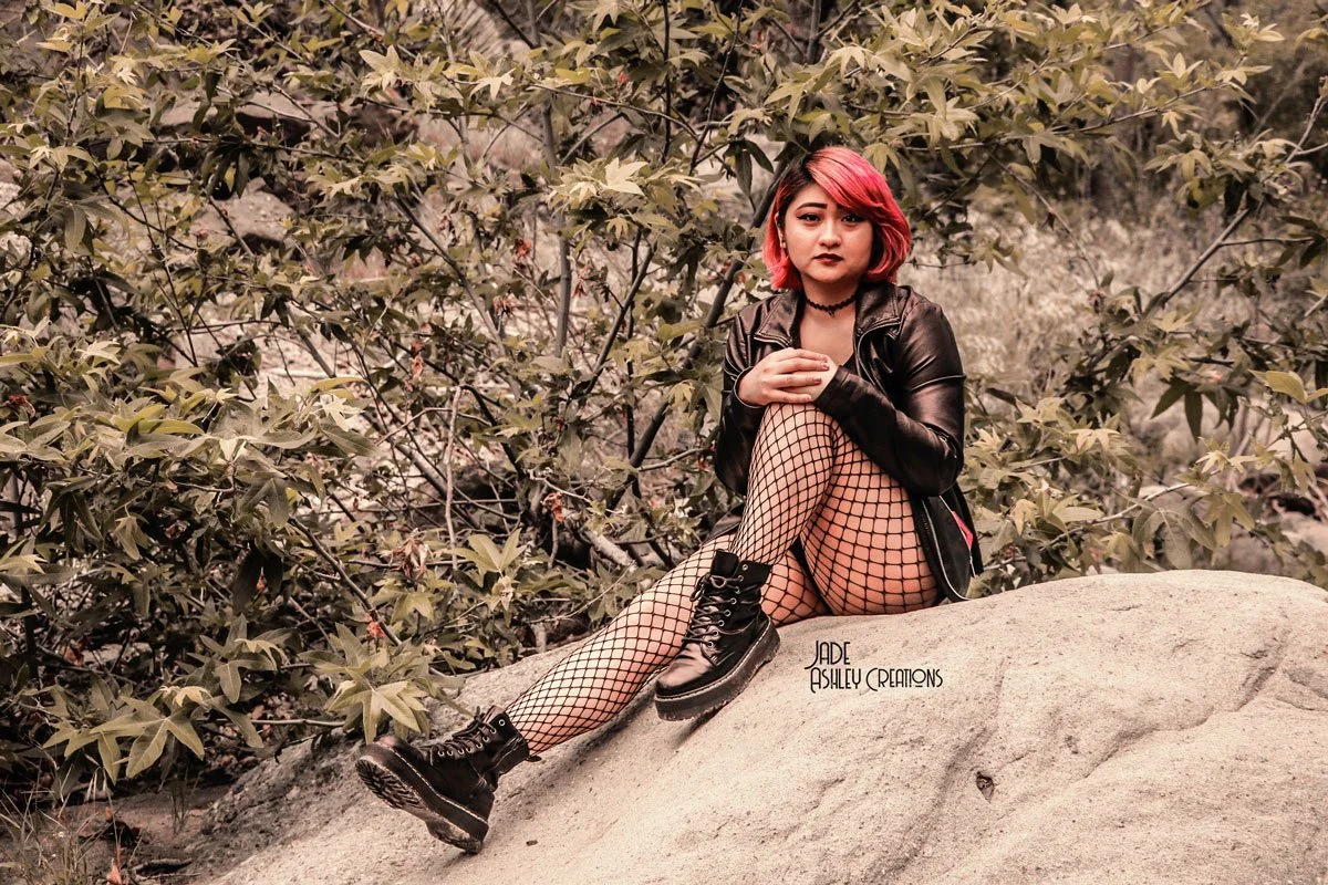 A young woman with pink hair sitting on a large rock surrounded by leaves and bushes, wearing a black leather jacket, fishnet stockings, and black combat boots.