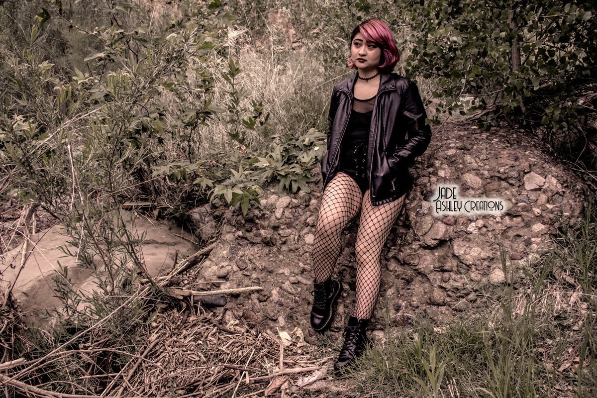 A young woman with pink and purple hair, wearing a black leather jacket, black dress, fishnet stockings, and black combat boots, sitting on rocks in a natural outdoor setting with green shrubs and dry grasses.