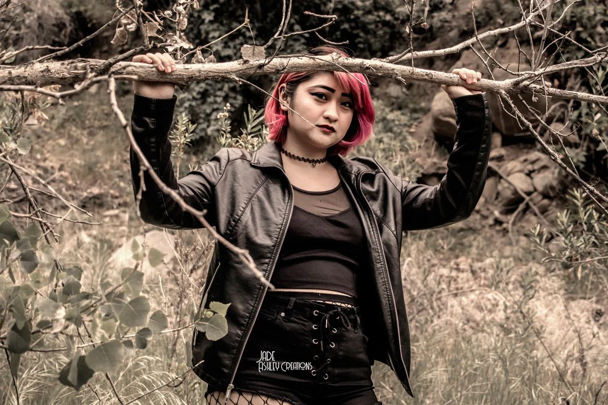 A young woman with pink hair wearing a black leather jacket, black crop top, and black shorts standing outdoors among branches and plants, holding a tree branch above her head.