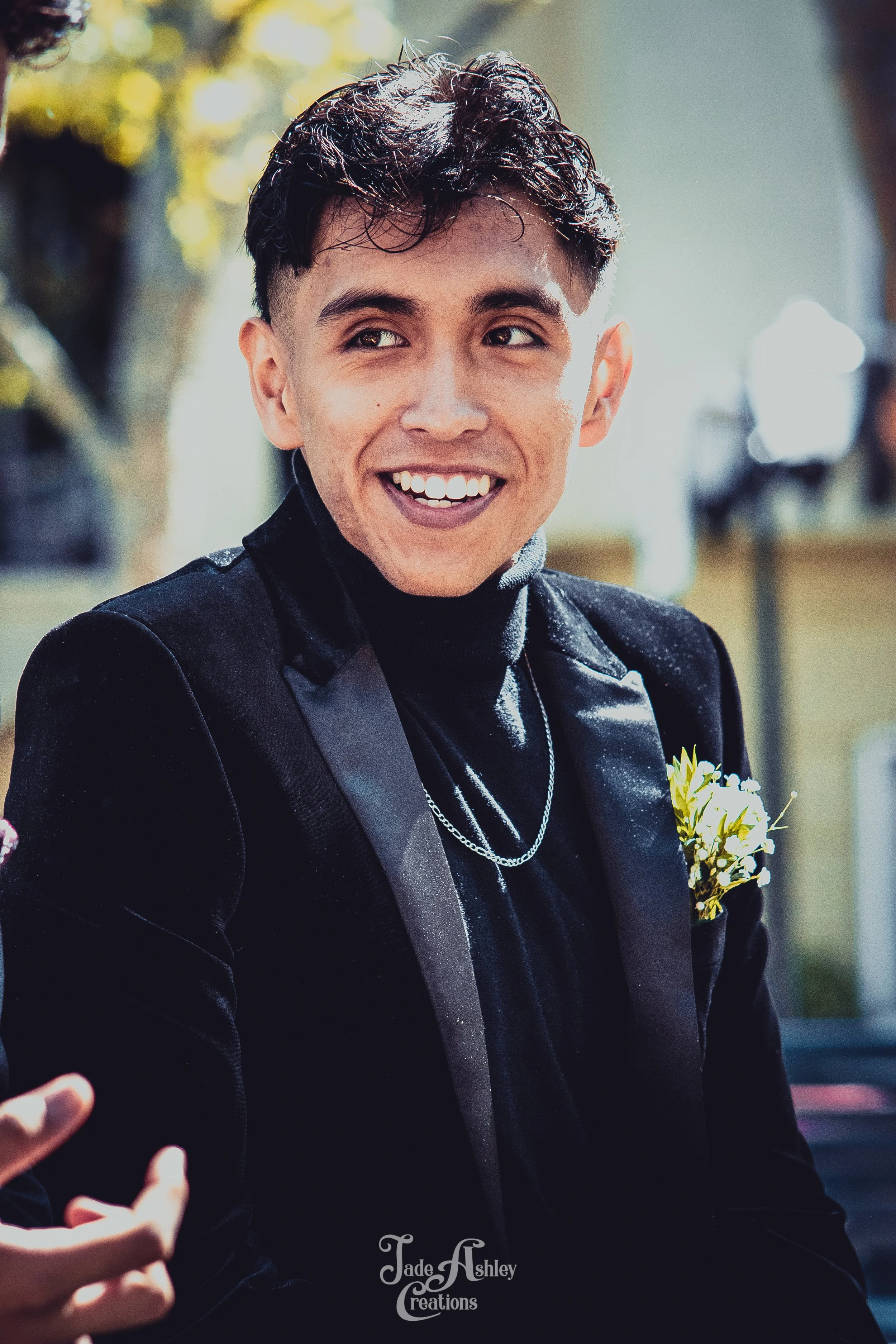 A young man dressed in a black velvet tuxedo, smiling outdoors during daytime, with a boutonniere on his chest.
