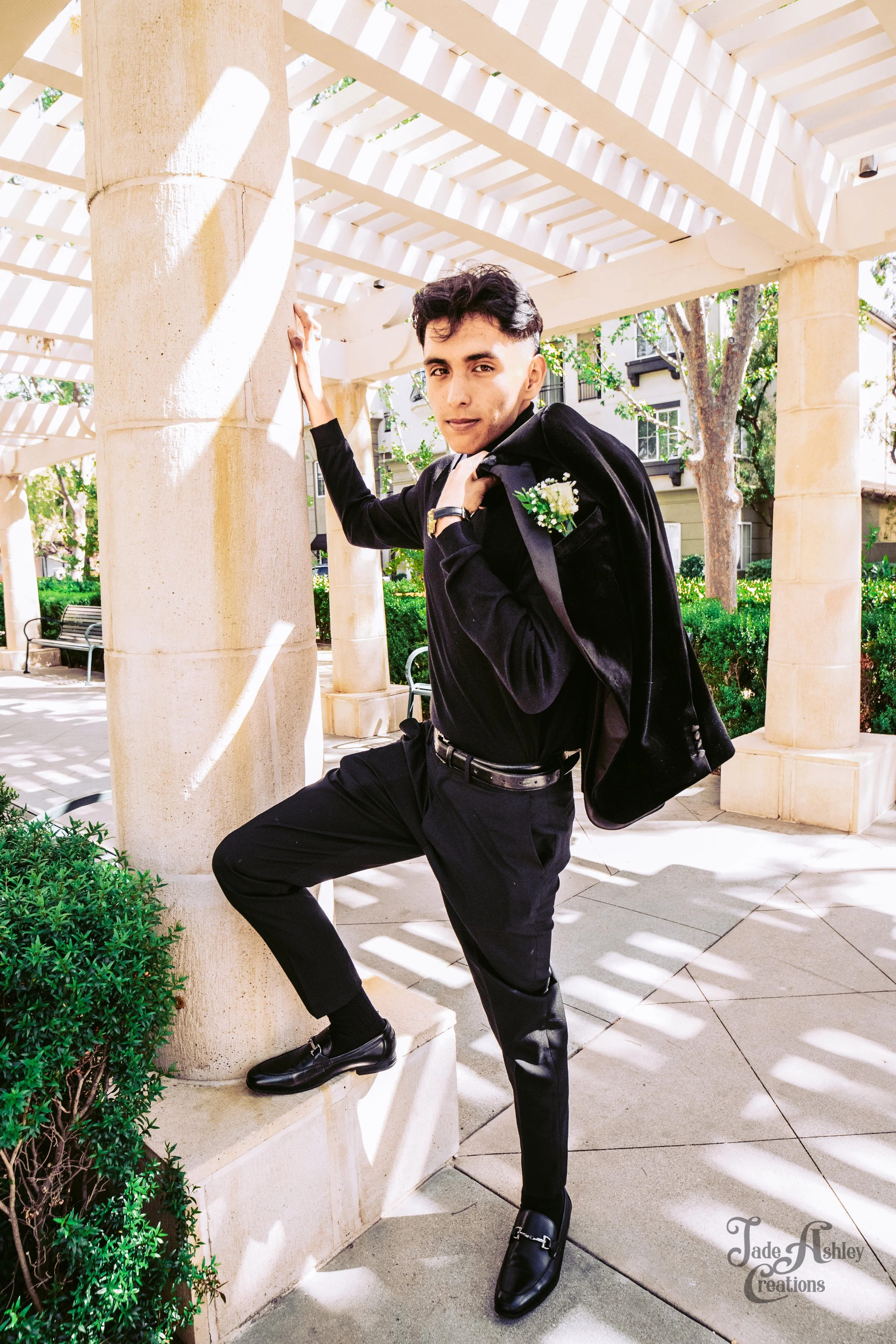 A young man in a black suit, with a boutonniere, poses with one foot on a stone base and one hand on a column in an outdoor area with a trellis and greenery.