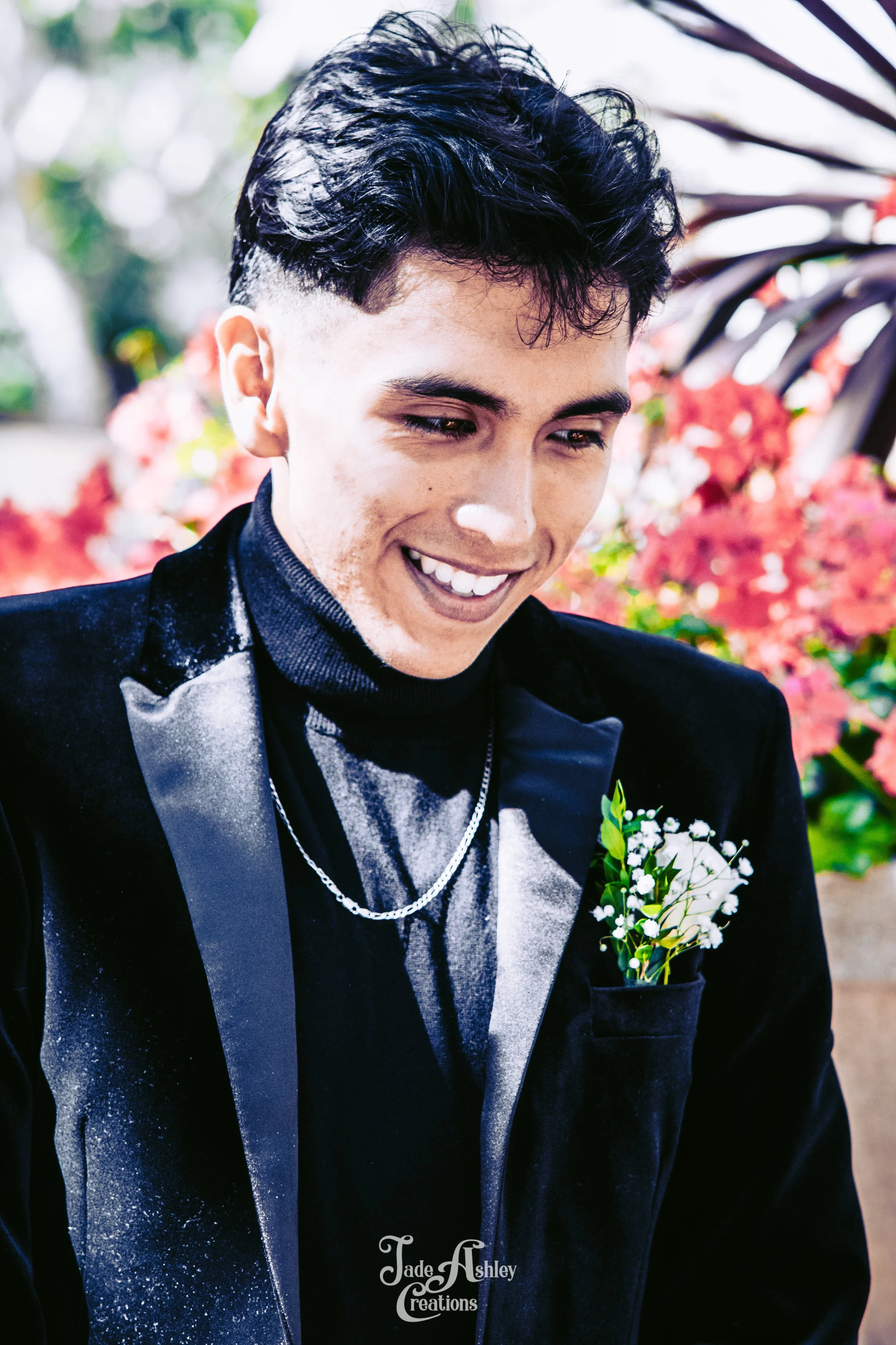 Young man smiling, wearing a black tuxedo with shiny lapel, black turtleneck, and a small white flower boutonniere on his jacket pocket, standing outdoors with pink and red flowers in the background.