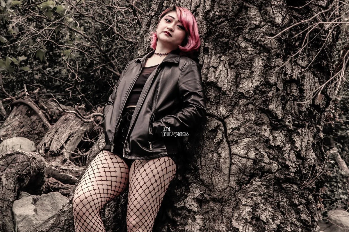 A woman with pink hair wearing a black leather jacket, fishnet stockings, and dark lipstick stands leaning against a large tree trunk in a natural outdoor setting with branches and leaves in the background.