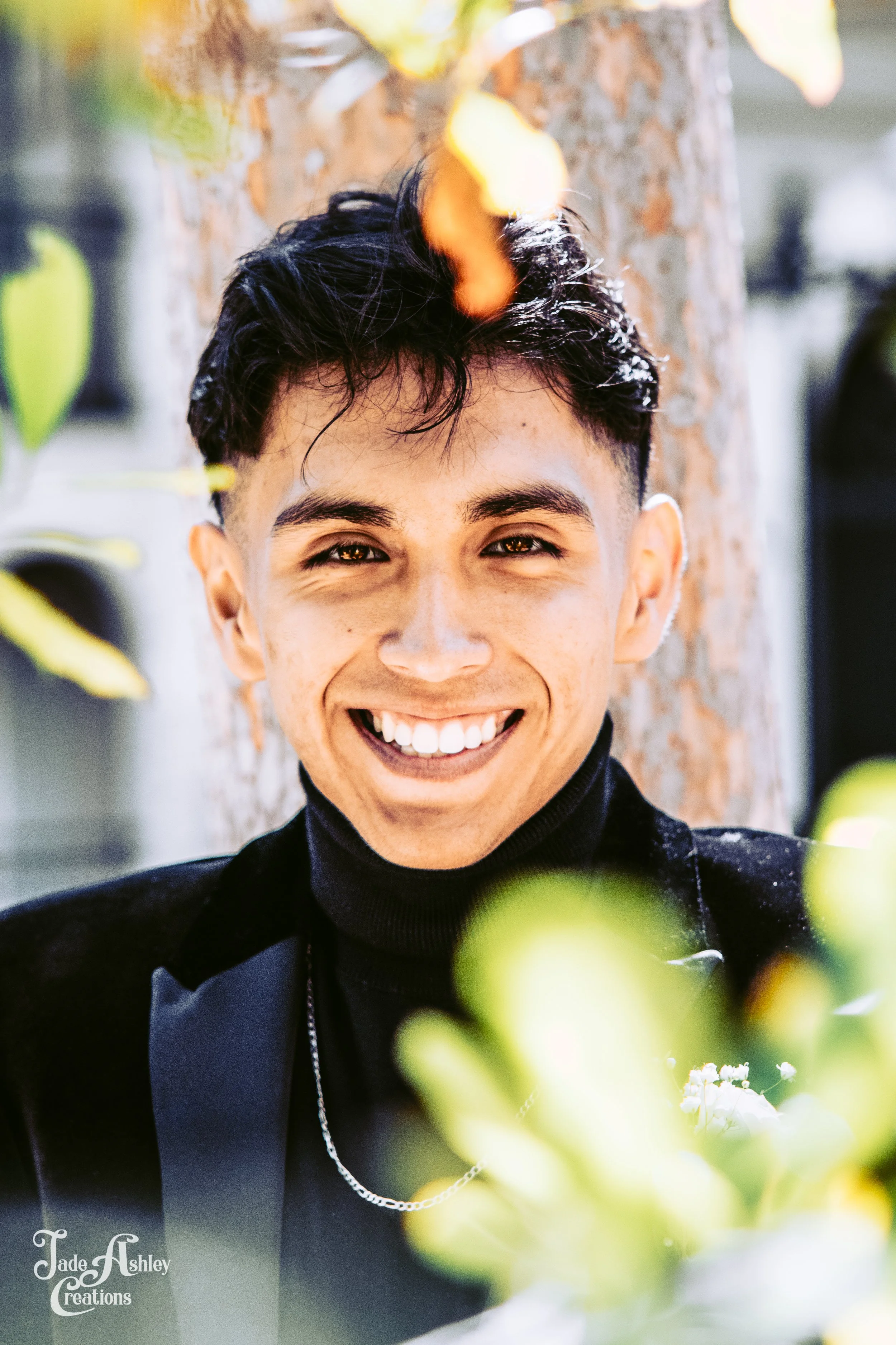 Smiling young man with short dark hair, wearing a black turtleneck and blazer, outdoors with tree and leaves in background.
