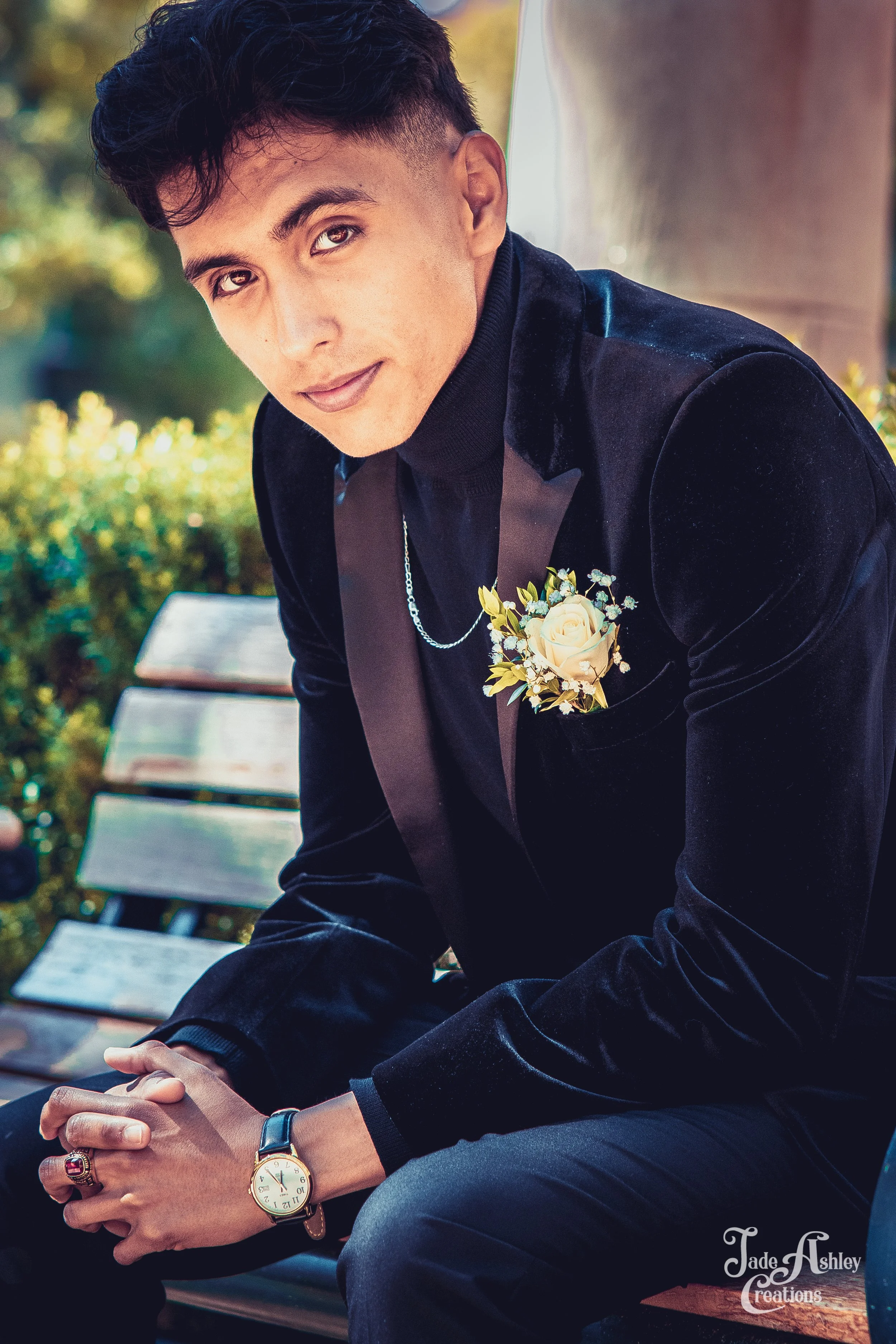 Young man dressed in a black velvet blazer with a boutonnière, sitting outdoors on a park bench, looking at the camera.